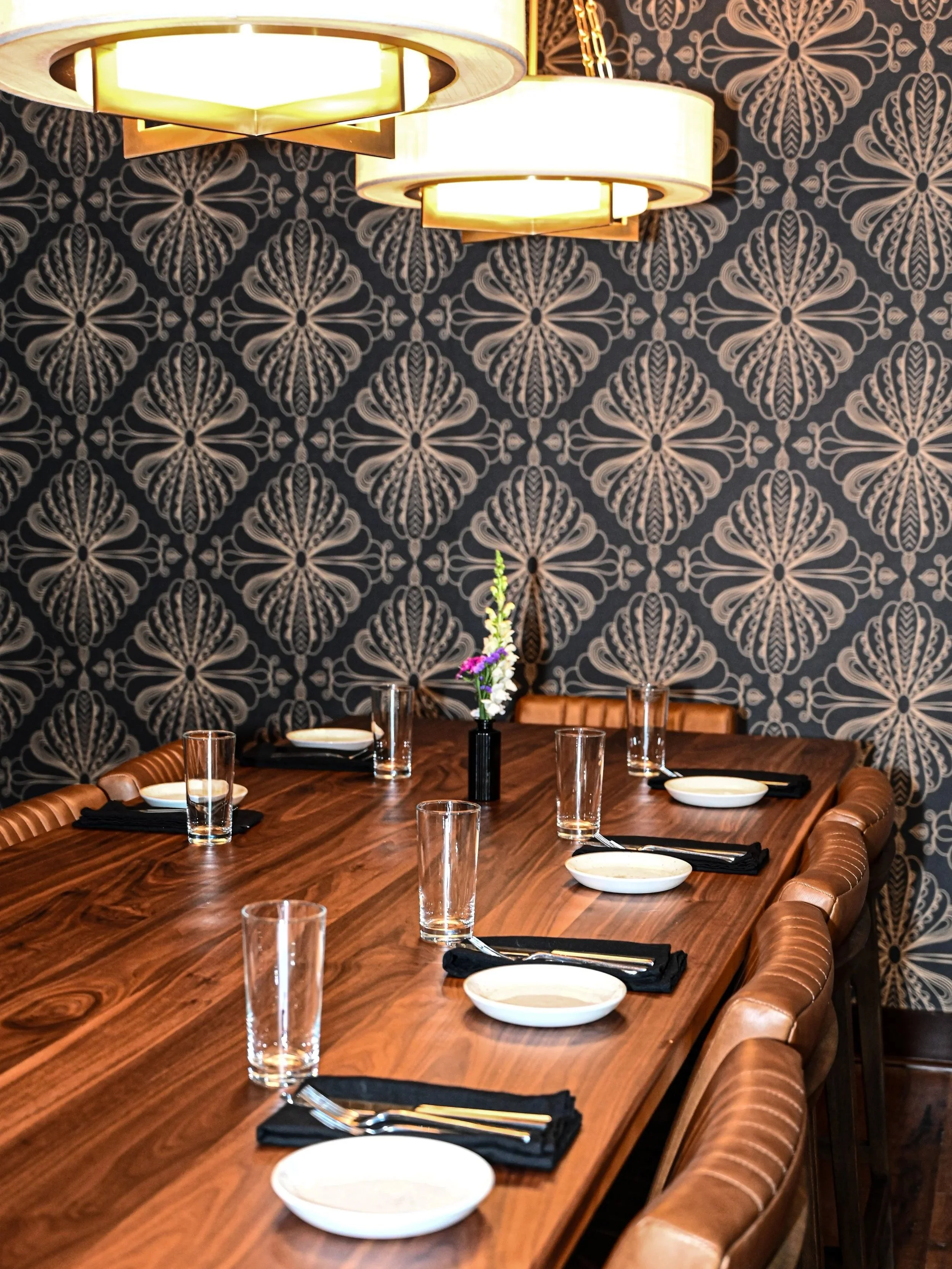 A dining room with a long wooden table set with white bowls, glasses, and black napkins, surrounded by leather chairs. The background features a dark wallpaper with a floral pattern, and modern lighting fixtures hang from the ceiling.