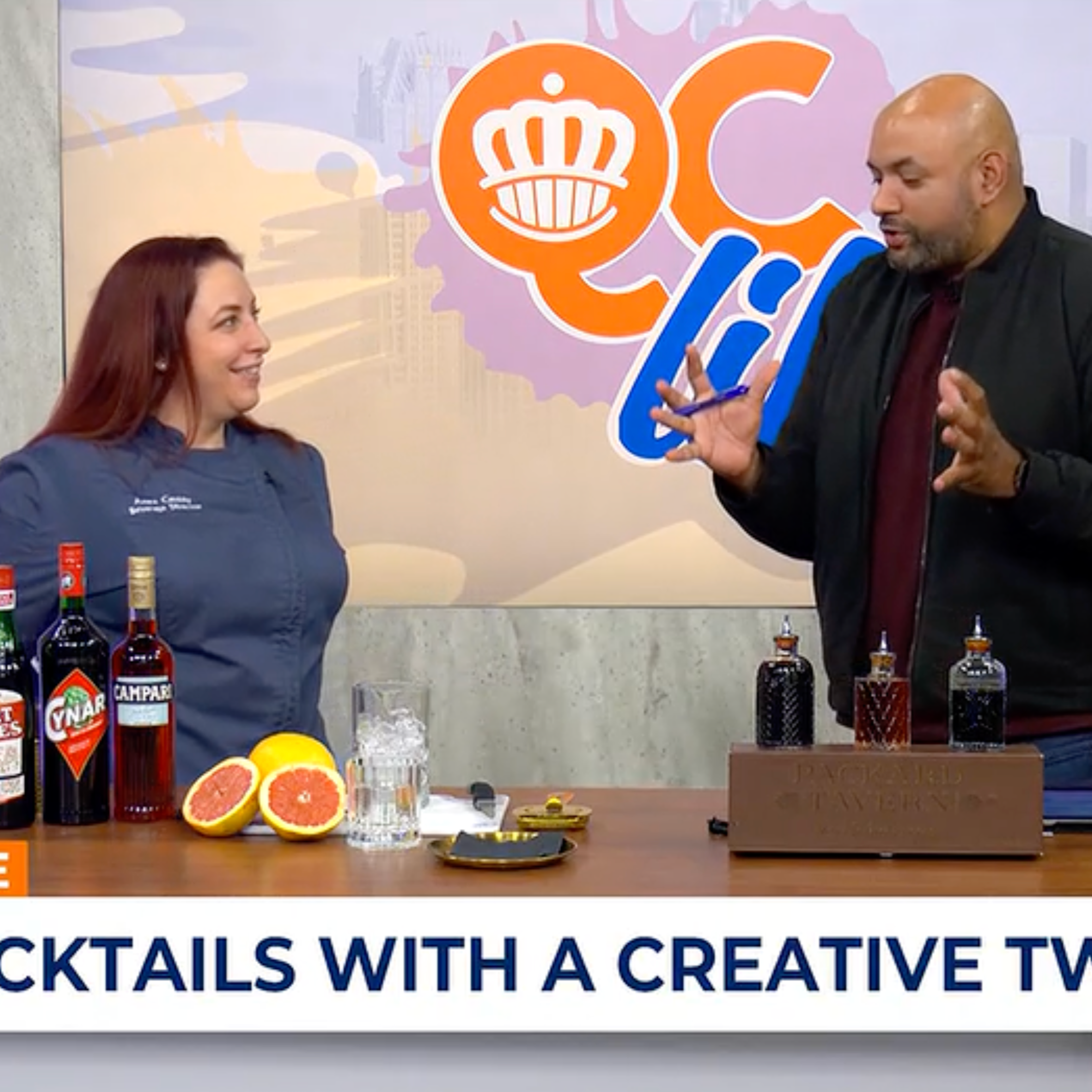 Two people engaging in a conversation near a table with various bottles and fresh grapefruit halves, with a backdrop featuring a colorful logo and cityscape illustrations.