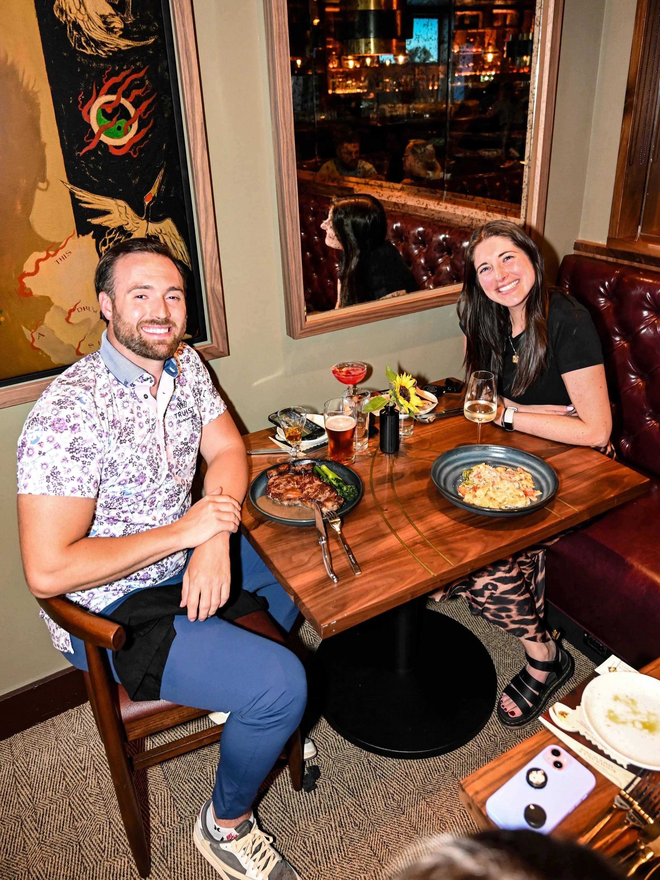 Two people sitting at a restaurant table with food and drinks, smiling at the camera. The person on the left has a beard and is wearing a floral shirt, and the person on the right has long dark hair, wearing a black top and black sandals. The table has a steak, pasta, wine, and cocktails. The background shows a window with a city view and some artwork on the wall.