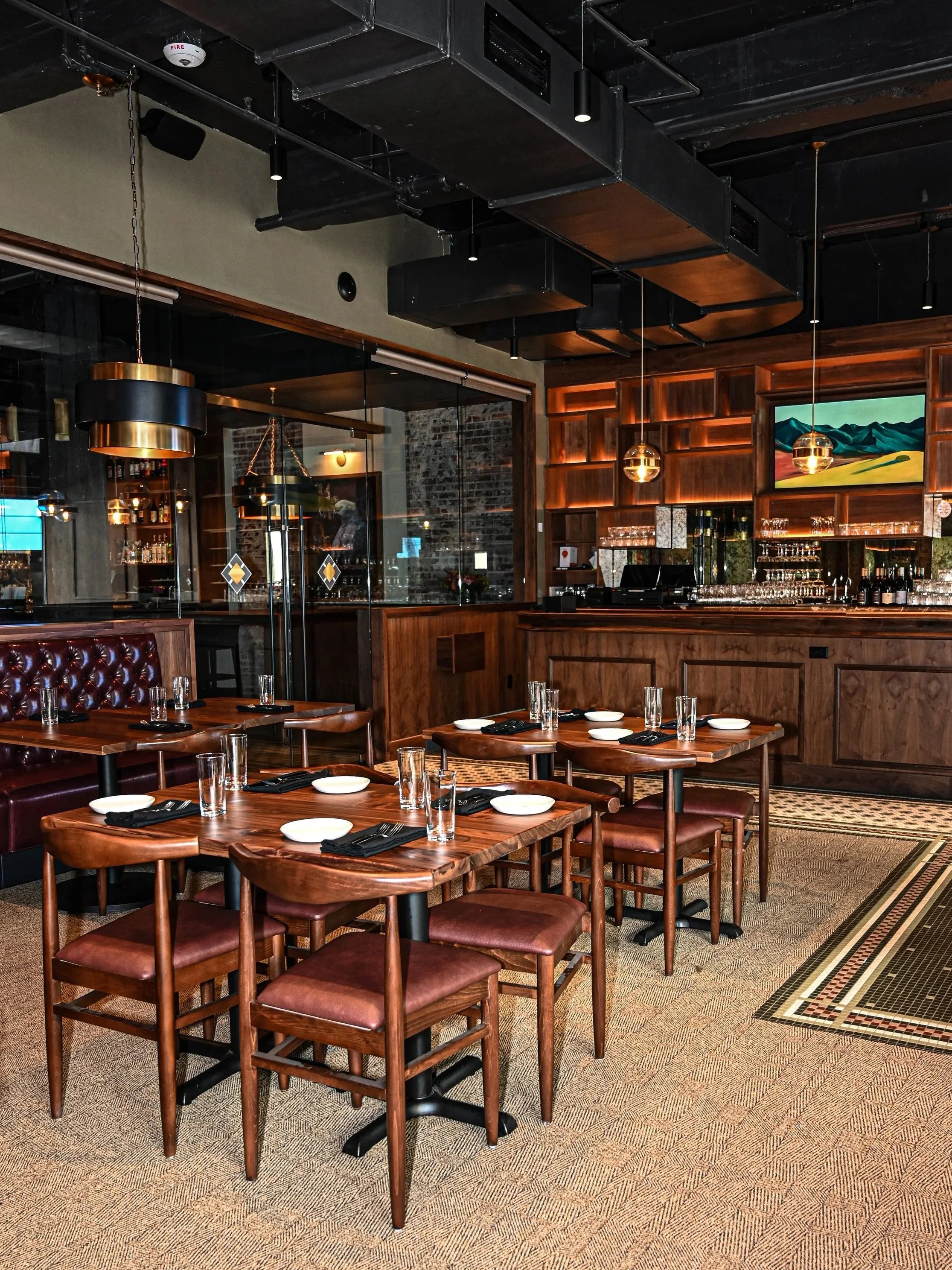 Interior of a restaurant with wooden tables and chairs, set with glasses, plates, and napkins, featuring a bar area with shelves of bottles and glasses, wooden wall paneling, and modern hanging light fixtures.