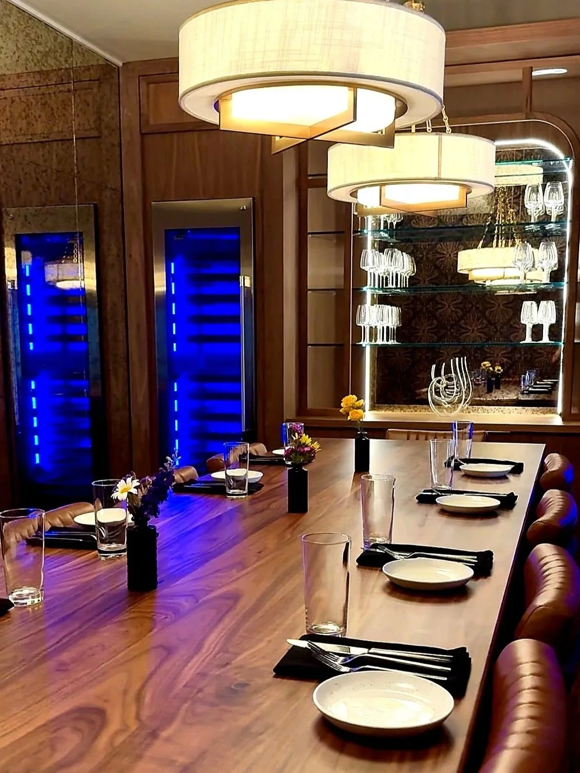 A modern dining room with a long wooden table set with plates, forks, and glasses, decorated with small vases of flowers. The room features wooden walls, a large light fixture hanging from the ceiling, and a mirrored wall with glass shelves displaying wine glasses and decorative items. There are two wine refrigerators with blue interior lighting.