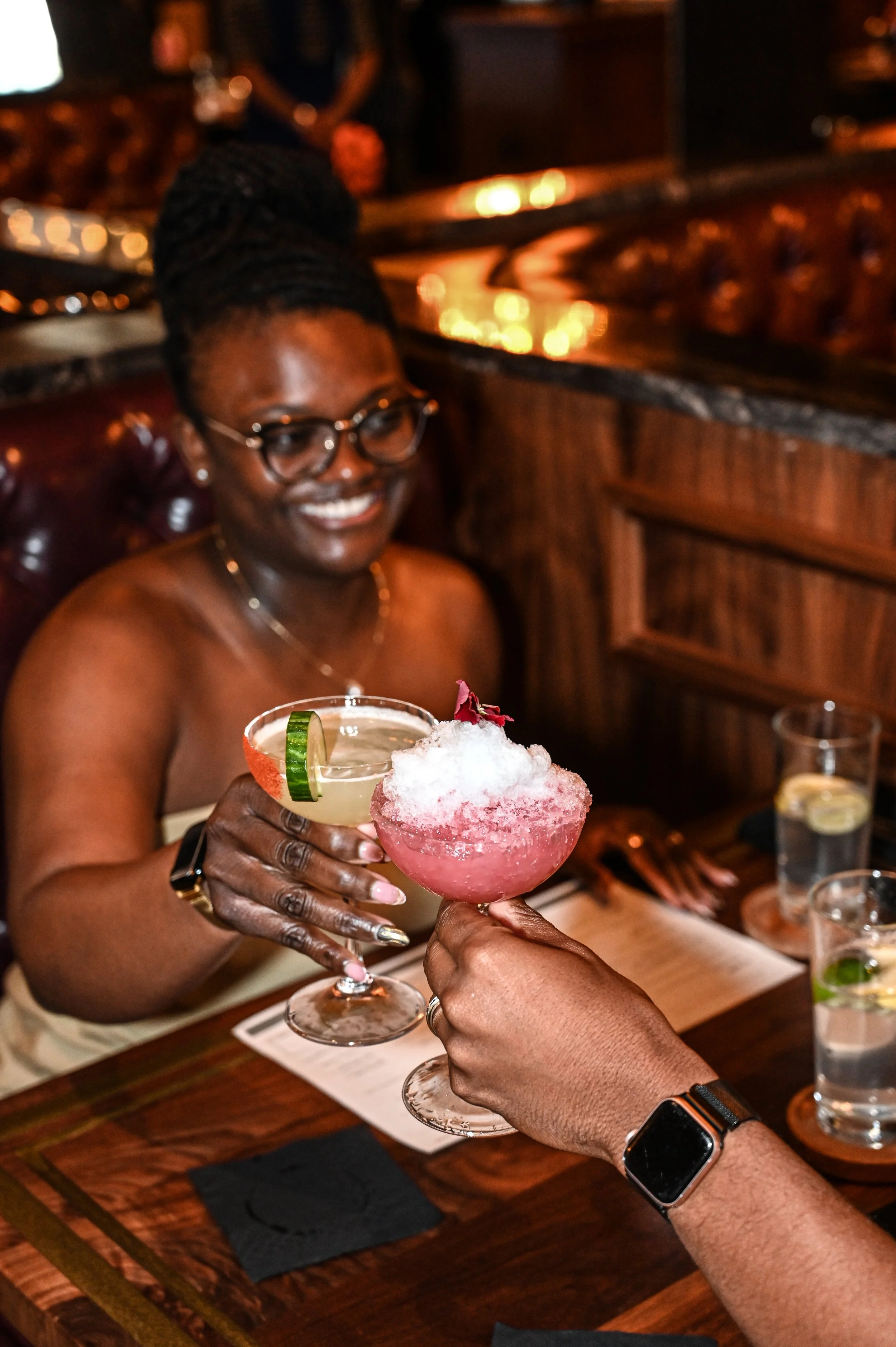 A woman with glasses and a braided hairstyle smiling while holding a pink frozen cocktail drink topped with shaved ice and garnished with a flower and lime wedge, at a restaurant or bar with warm lighting.