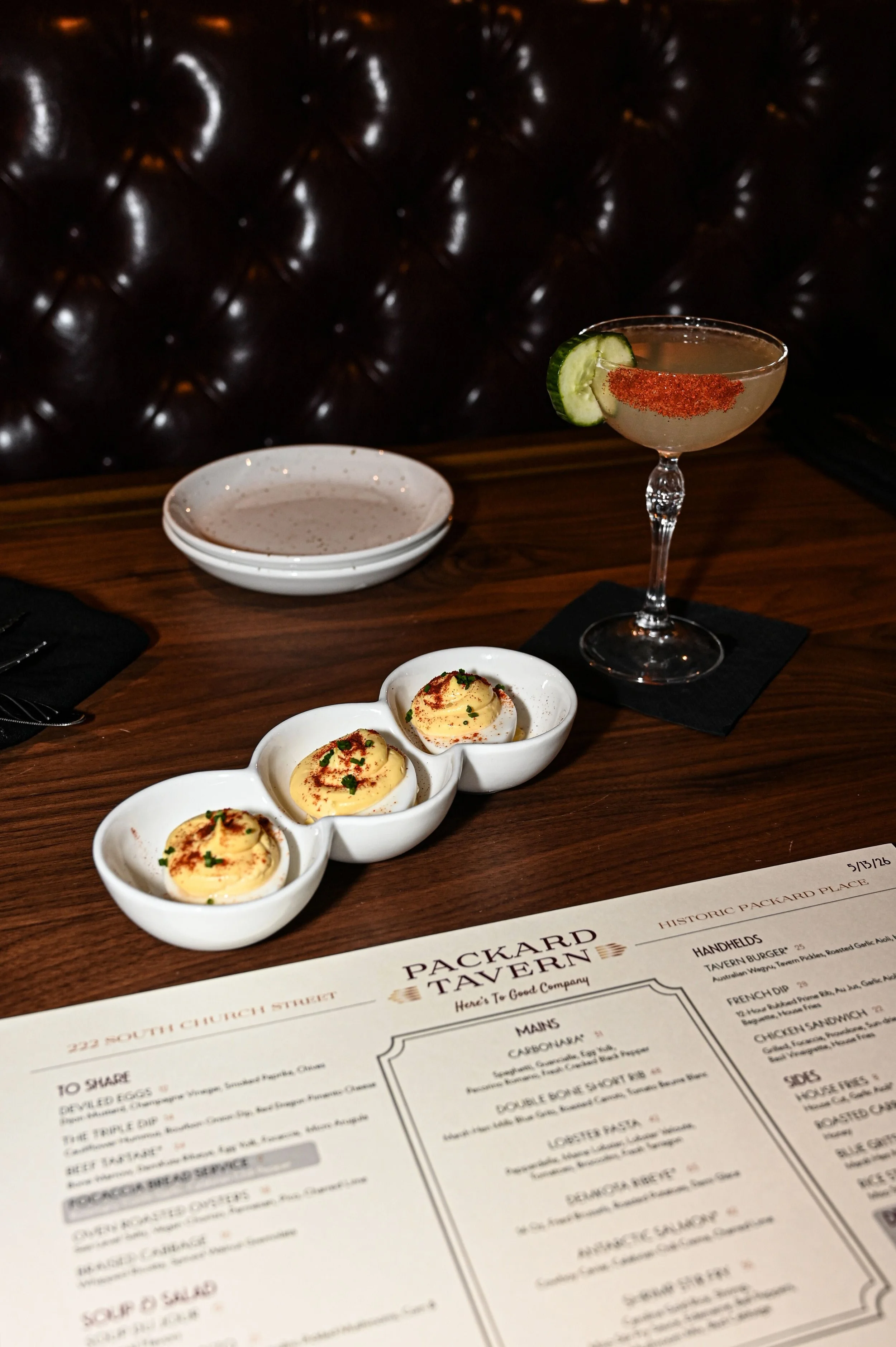 A restaurant table with a bar menu, a plate of deviled eggs garnished with paprika and chives, and a cocktail garnished with a pickle slice and chili powder, against a dark, tufted leather background.