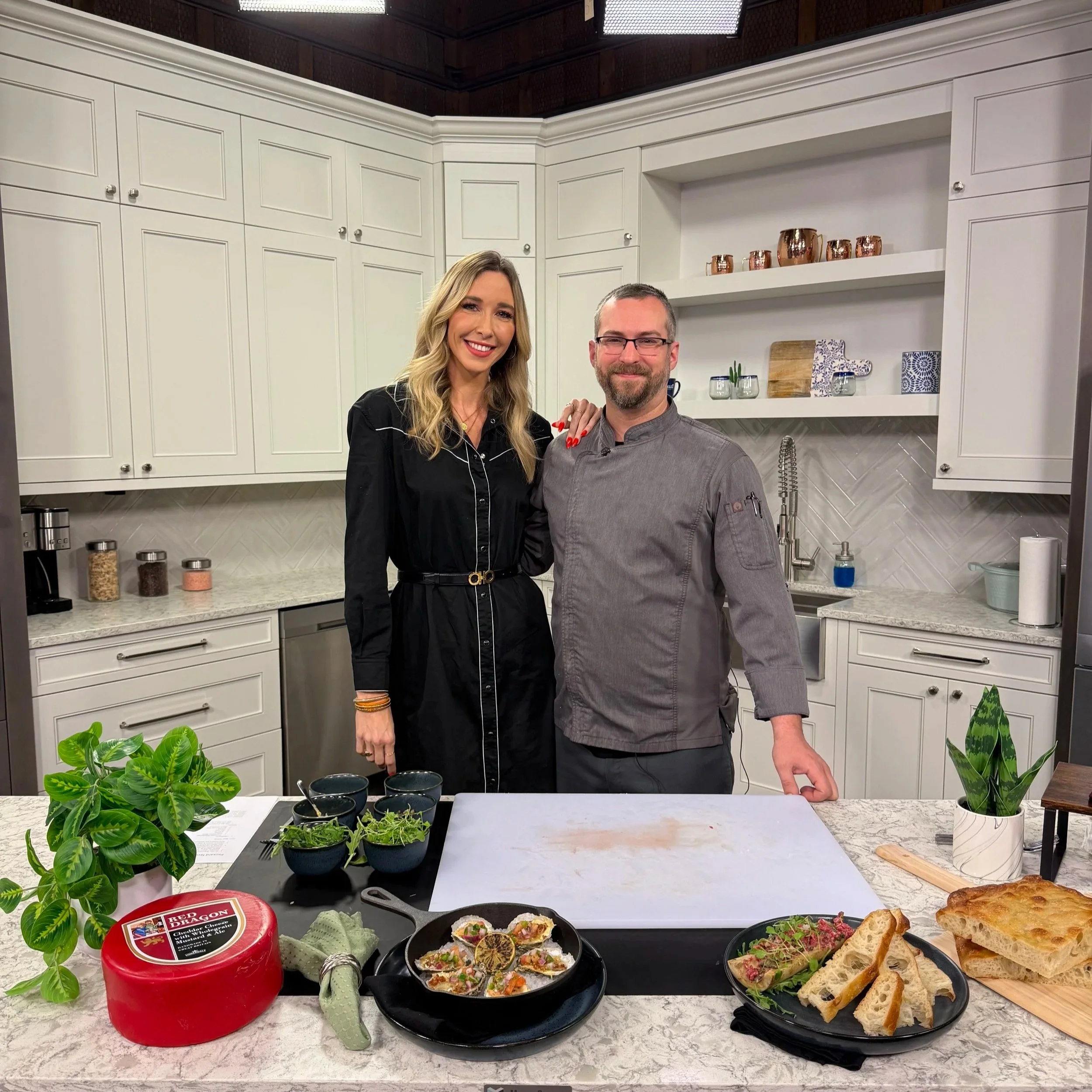 A woman and a man smiling in a kitchen with white cabinets. The kitchen counter has various food items, including bread, dips, and plants, and pots and copper cups are on the shelves behind them.