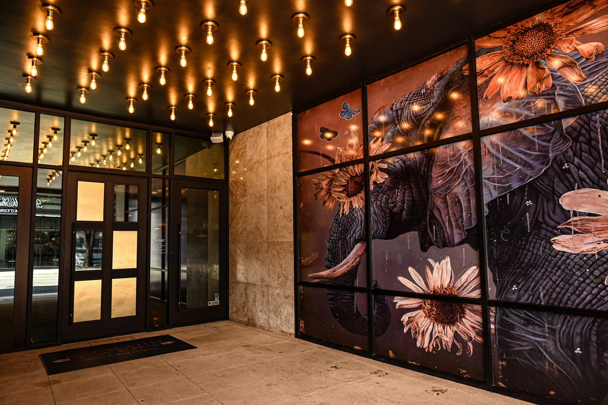 Lobby entrance with glass doors and a large mural of a dragon intertwined with flowers and butterflies on the wall. Modern lighting fixtures with exposed bulbs hang from the dark ceiling.