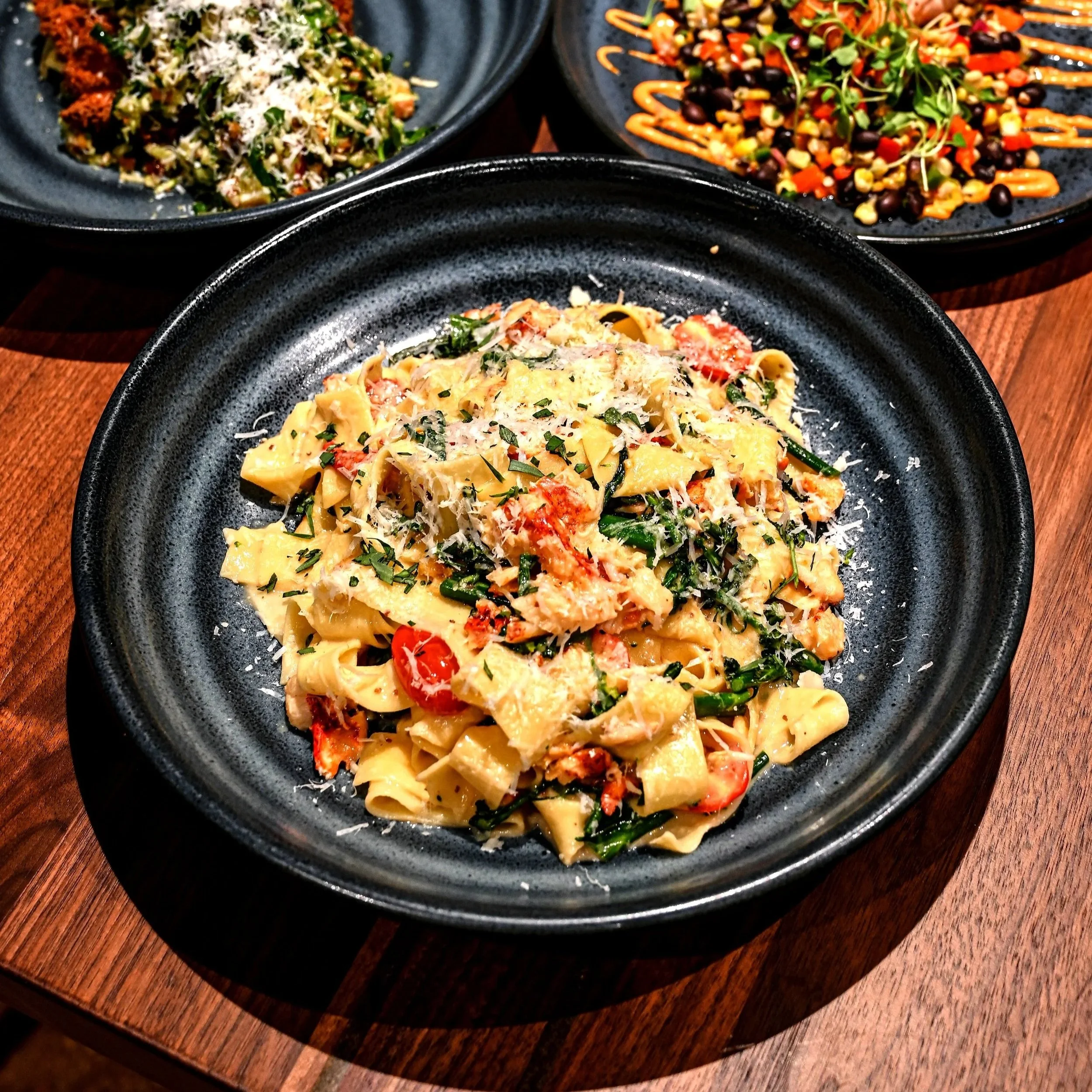 Three black plates of colorful pasta salads on a wooden table.