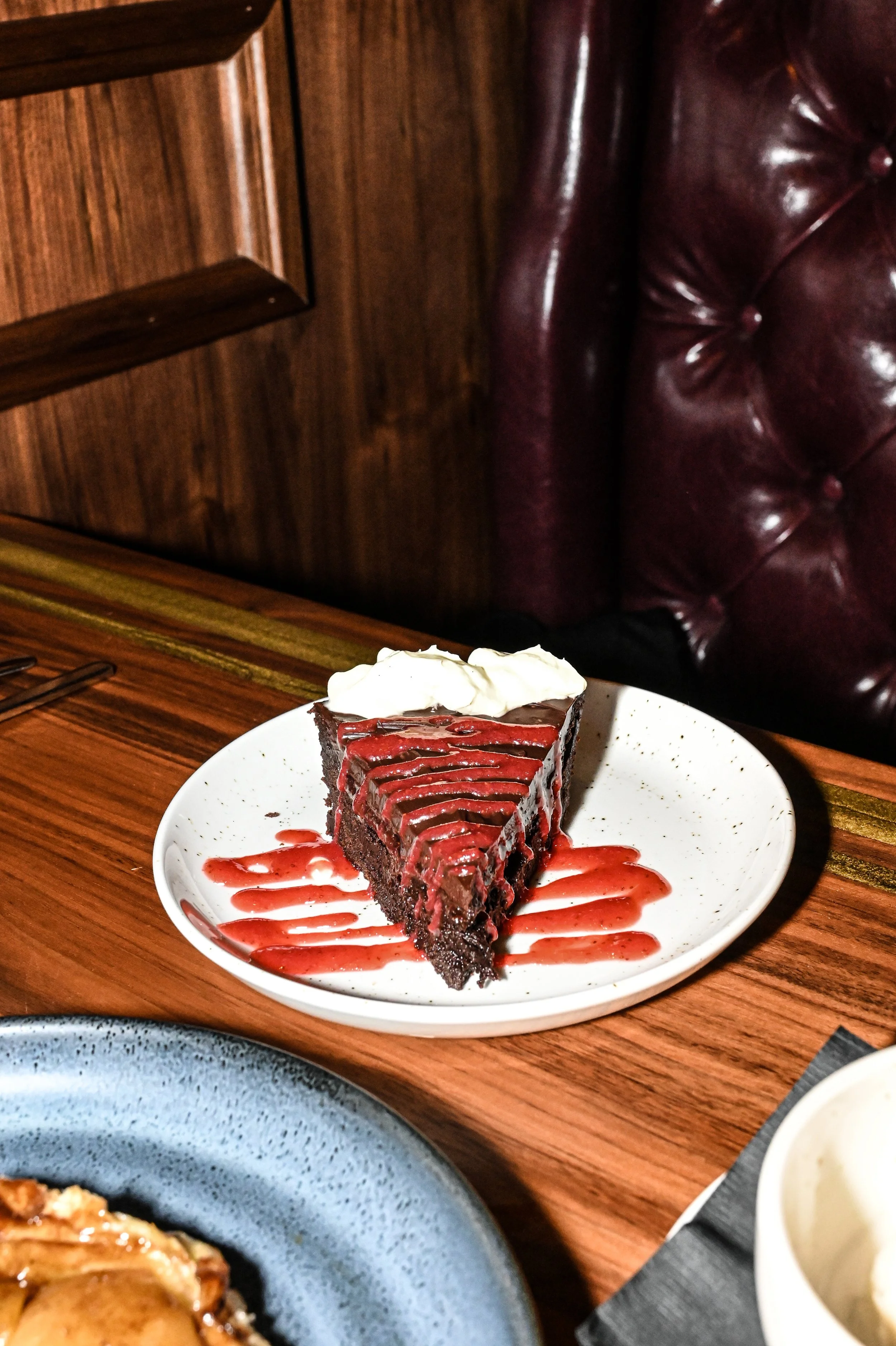 A slice of chocolate cake with whipped cream topping on a white plate, drizzled with red fruit sauce, placed on a wooden table in a restaurant setting.
