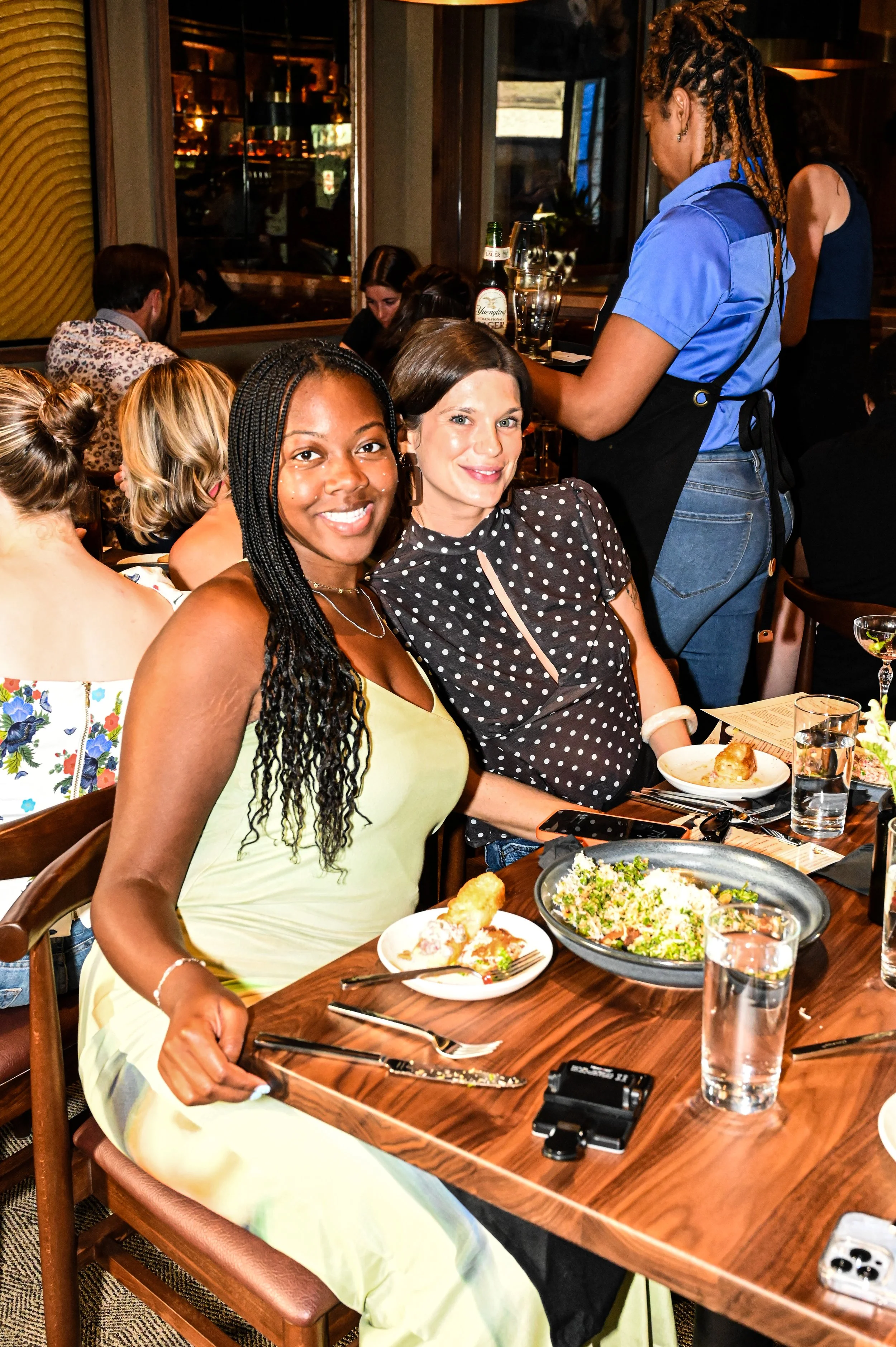 Two women sitting at a restaurant table with plates of food and salads, smiling at the camera. A waitress stands nearby, and other diners are visible in the background.