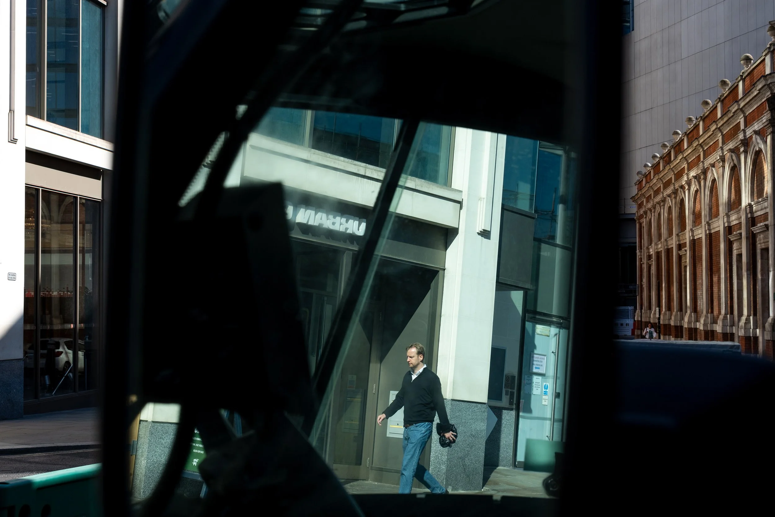 A city street scene viewed through a backview mirror; a man in a dark jacket and light pants is walking, holding a bag in his right hand; modern buildings with glass facades and a historic brick building with arched windows are visible.