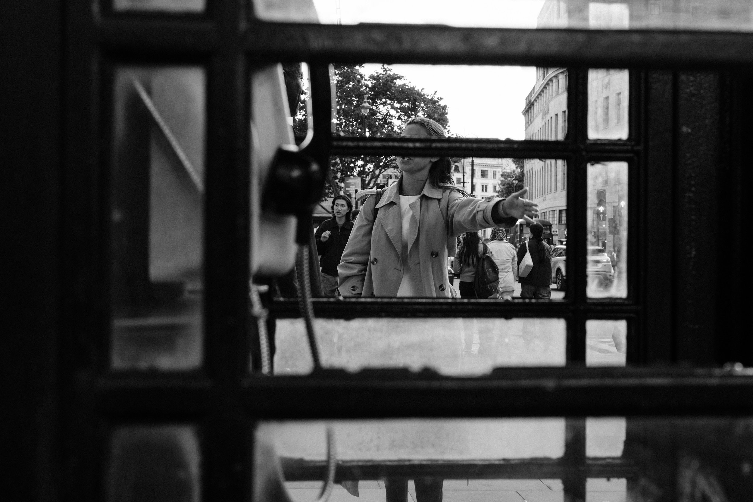 A woman standing behind a phone both, gesturing. Her face is partially obscured by a window beam of the phone booth.