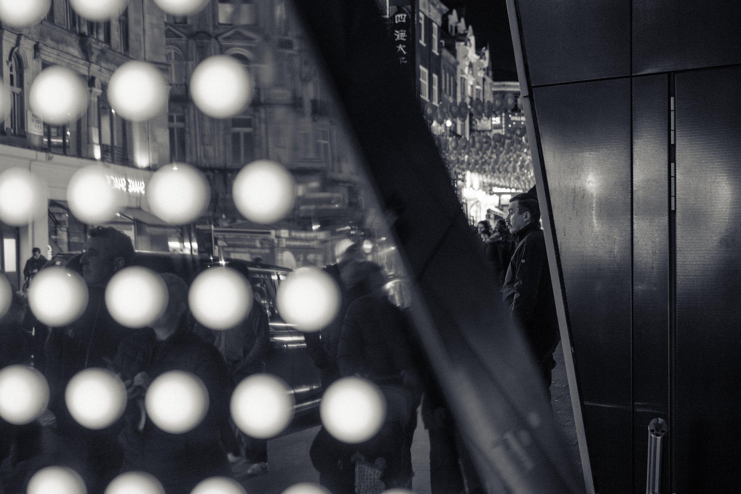A layered photograph shows a reflection of city buildings and street scene through a glass panel with a grid of illuminated white dots, and a person with short hair and a dark jacket standing outside.