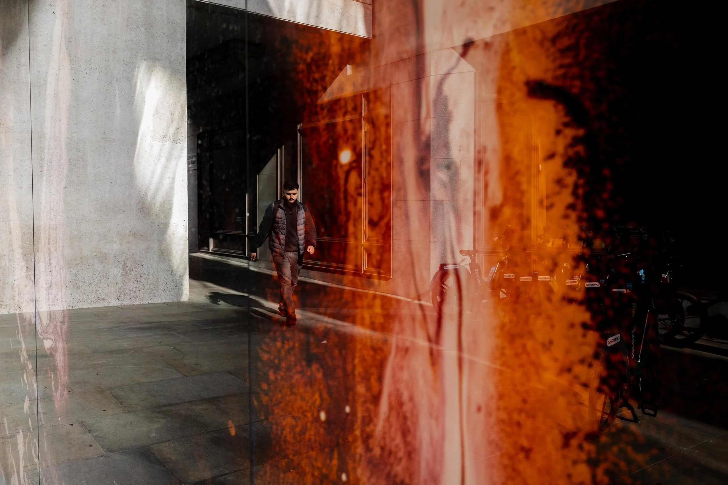 A man in a black vest and gray pants walking outside a building with large windows. The reflection and orange tint on the glass create an abstract pattern.