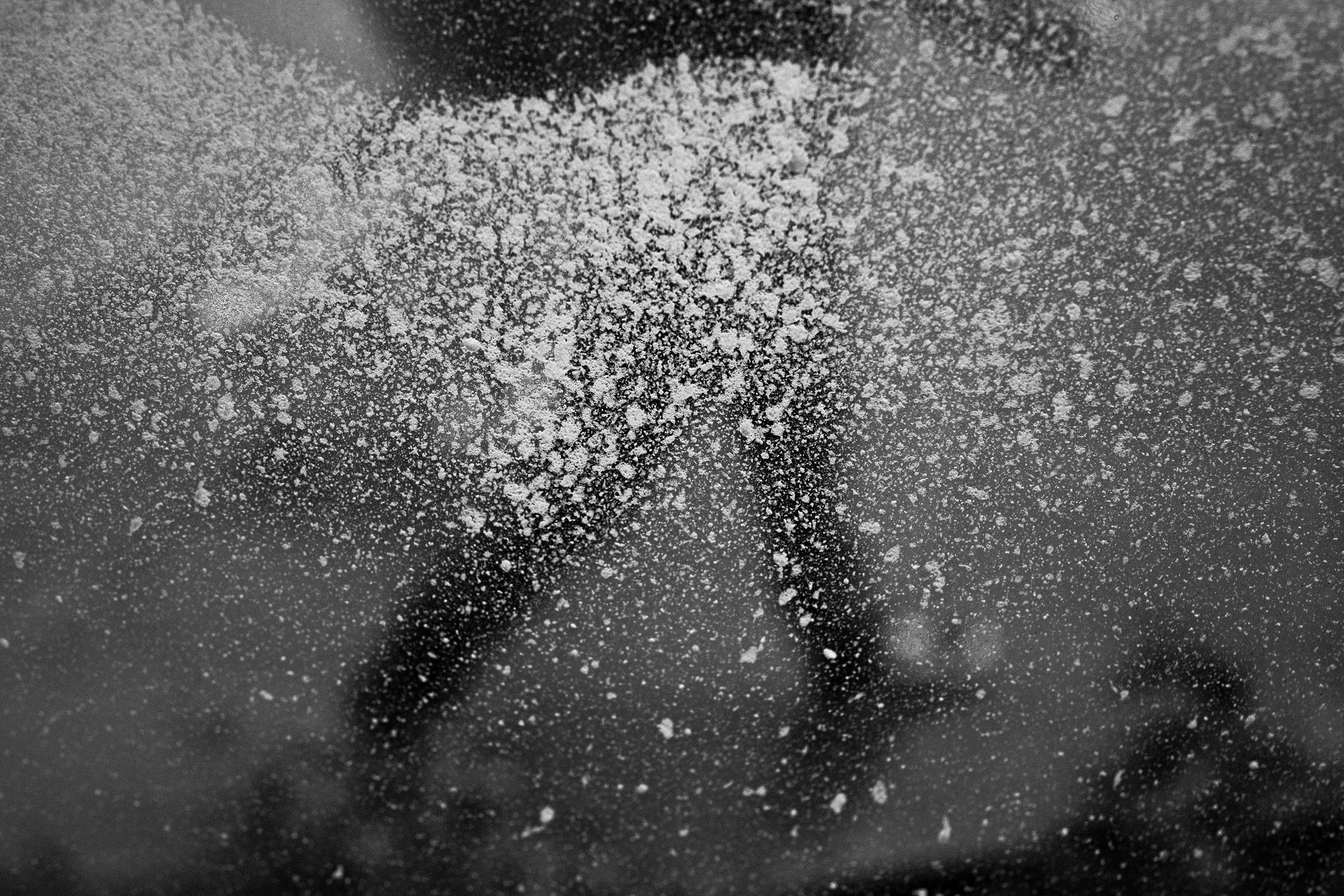 Close-up of a surface with scattered white powder resembling flour or powdered sugar and a partially visible black object in the background.