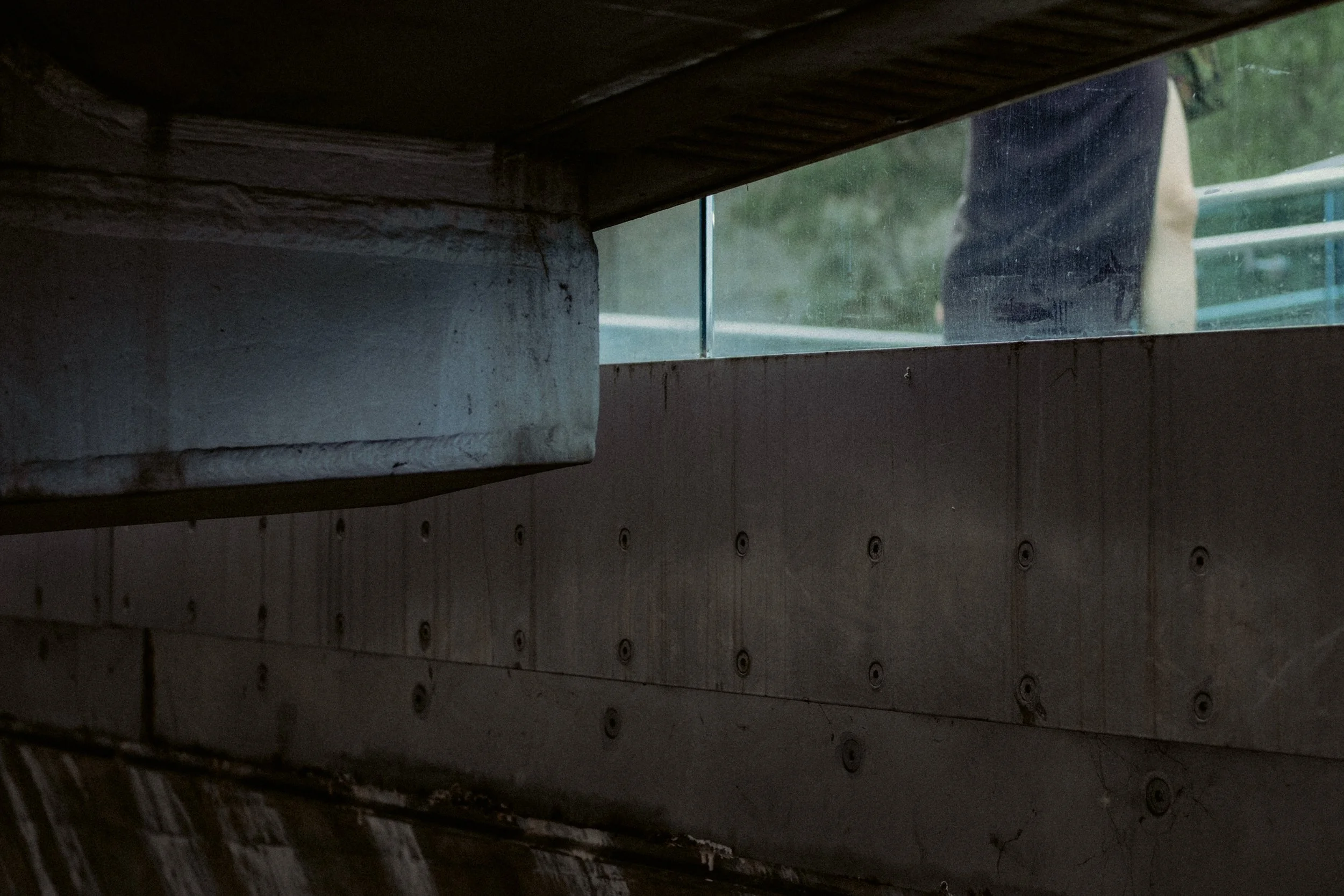 A view through a narrow rectangular opening under a bridge showing a person wearing dark clothing and white shoes on a bridge with green railings.