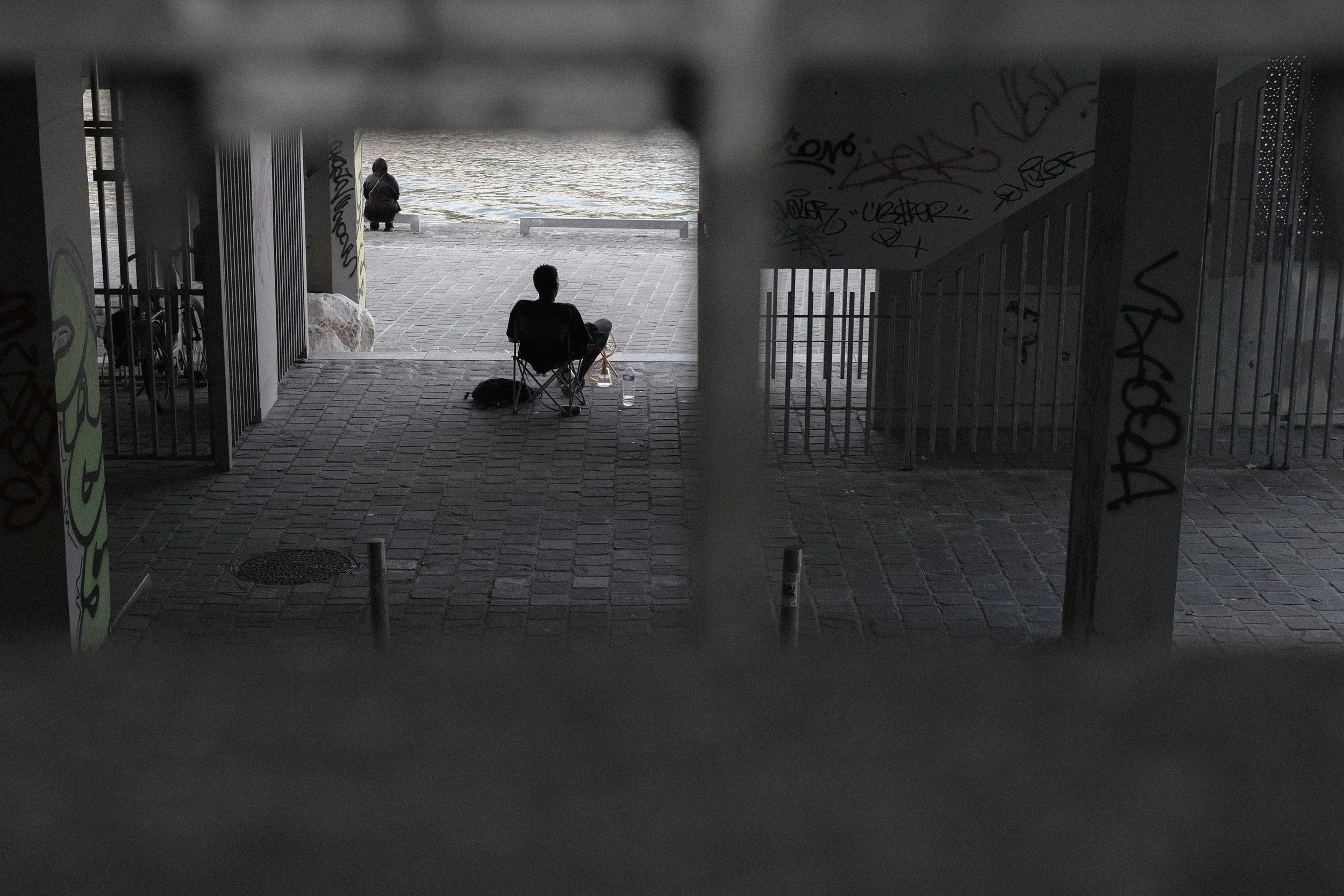 A view from inside a dark underpass looking out towards a waterfront. Two people are visible: one sitting on a folding chair with water bottles, and another standing close to the water's edge, dressed in a dark jacket. The scene is gloomy, with graff
