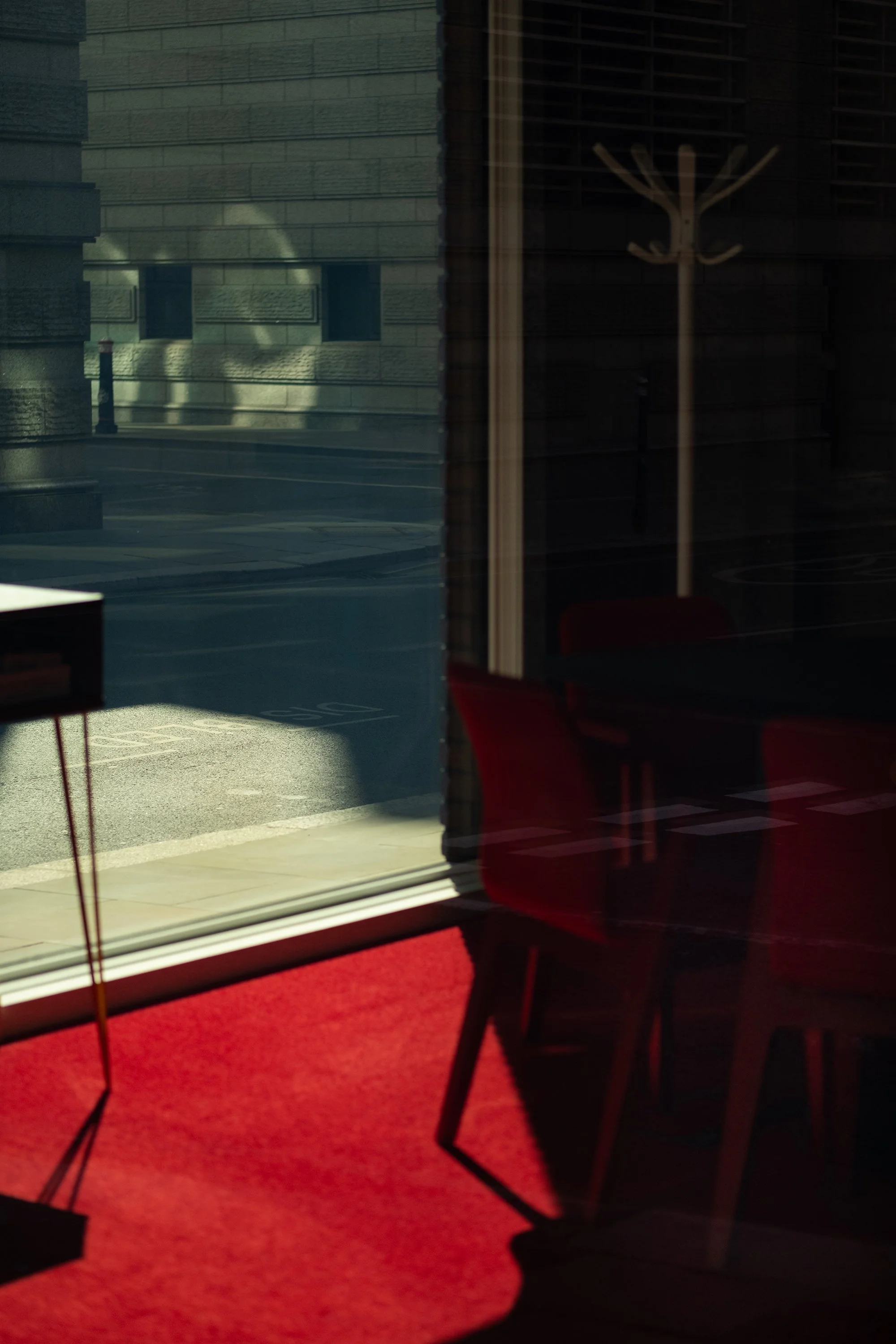A view through a glass window showing reflection of building facades, a street, and a parking space outside. Inside, red chairs and a table are visible, with sunlight casting shadows on the red carpet.