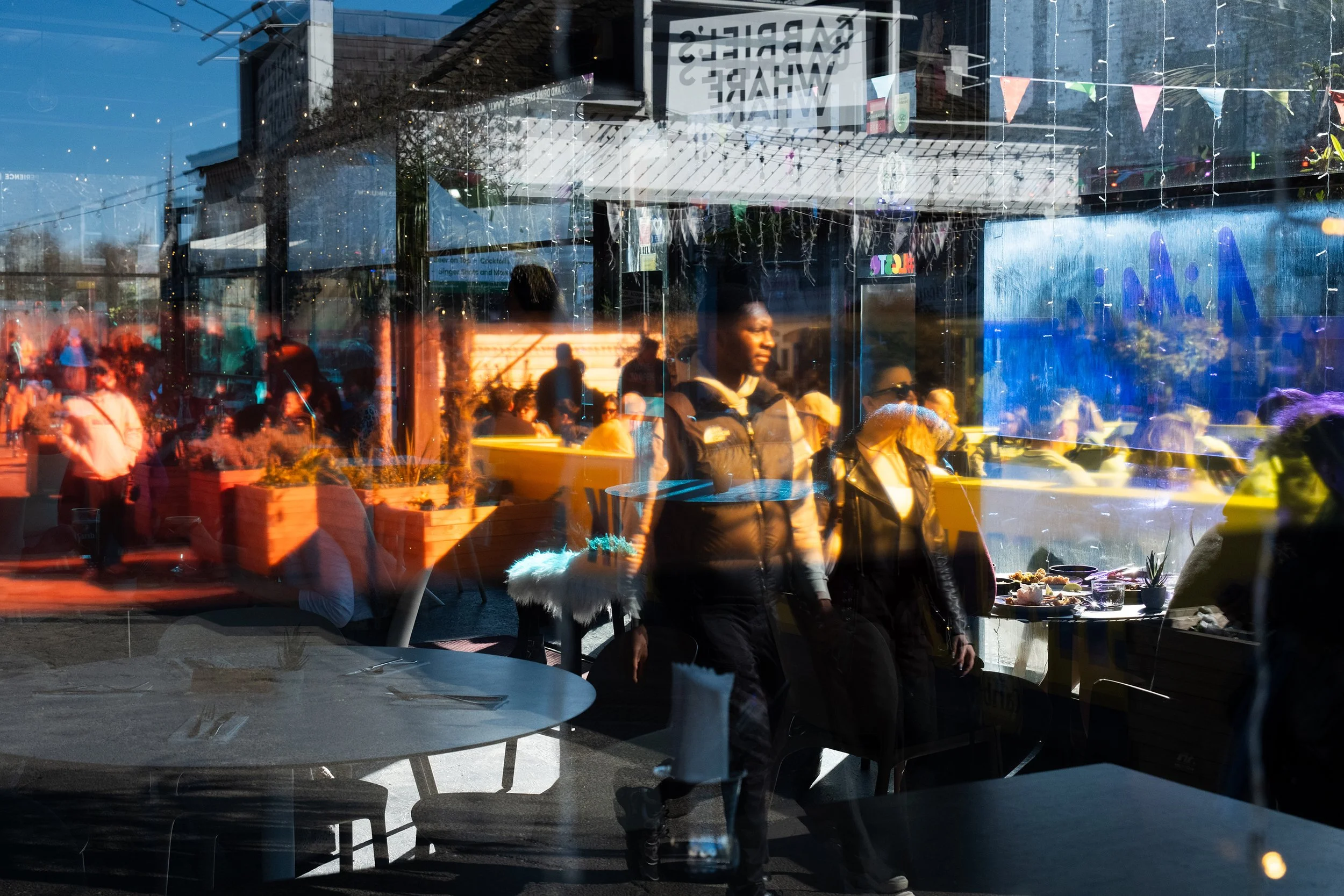 Reflected image of a restaurant window showing people dining outside, with some tables and food visible inside, and a 'CARRIER'S WAFFLE' sign, sunlight illuminating the scene.