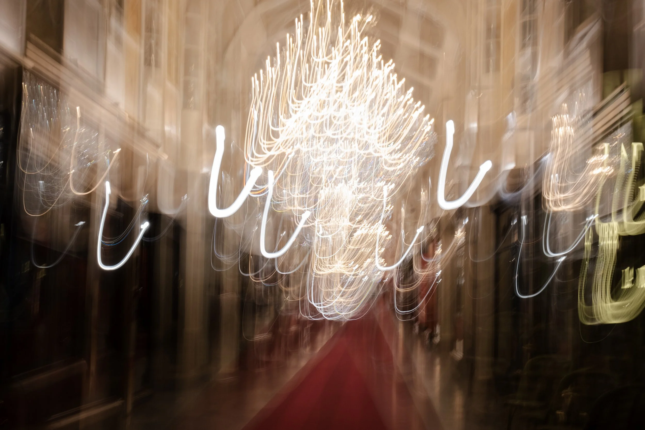 Blurred image of a chandelier with multiple lights in a hallway or entrance of a building, with a red carpet on the floor.