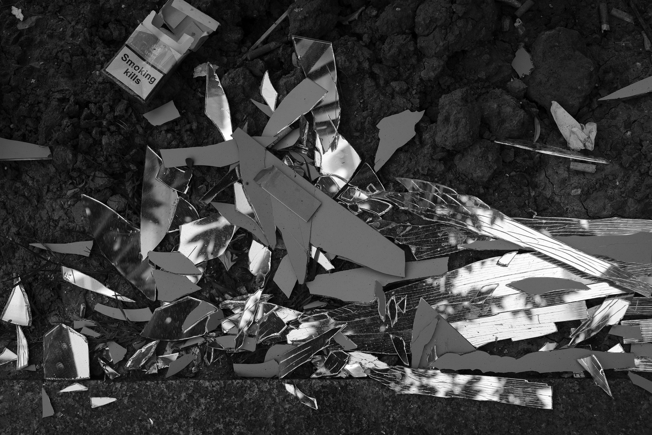 Broken mirror shards scattered on dark dirt ground, with a cigarette pack labeled 'Smoking kills' nearby.