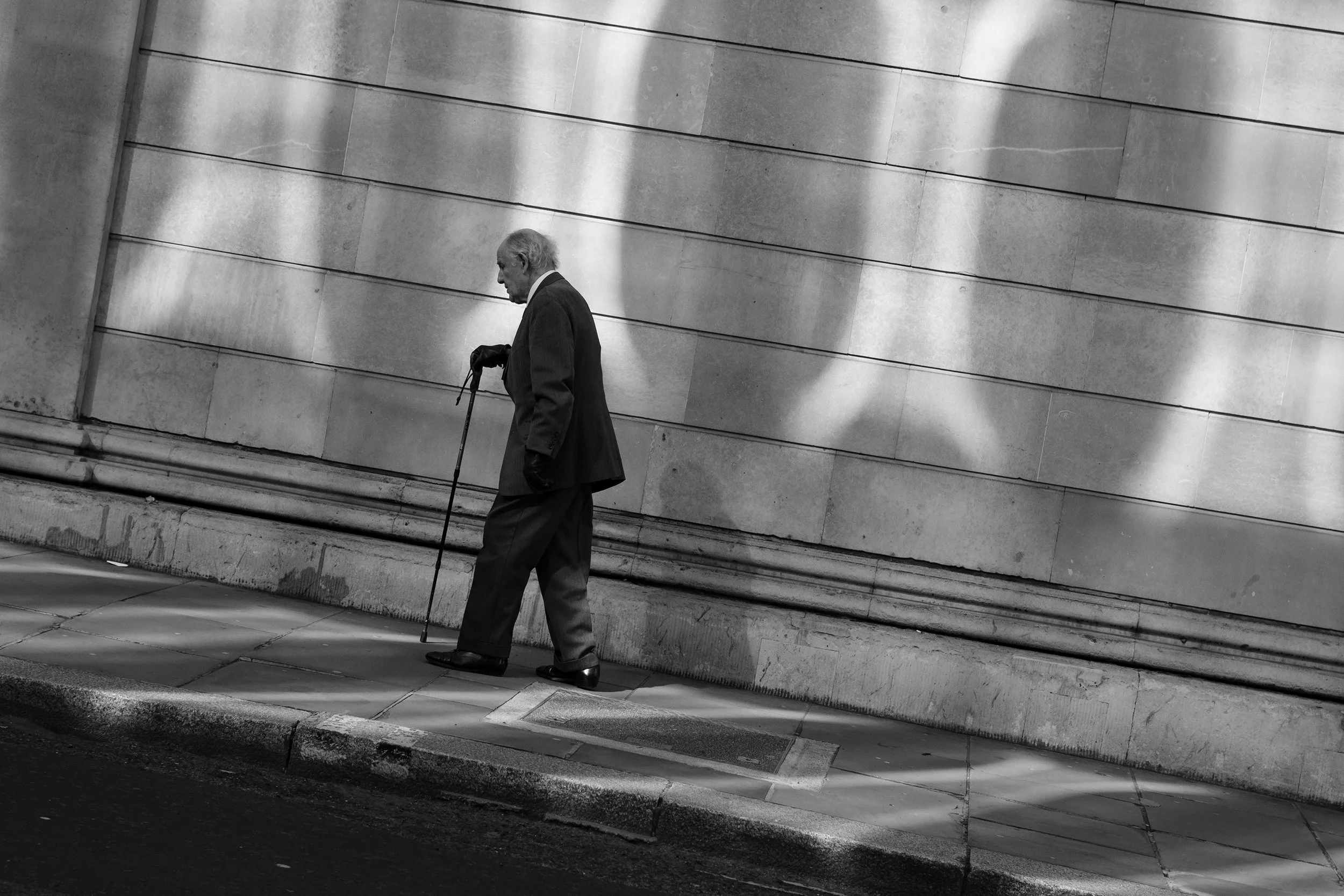 A black and white photo of an elderly man walking with a cane along a sidewalk beside a large stone building, casting a shadow on the wall.