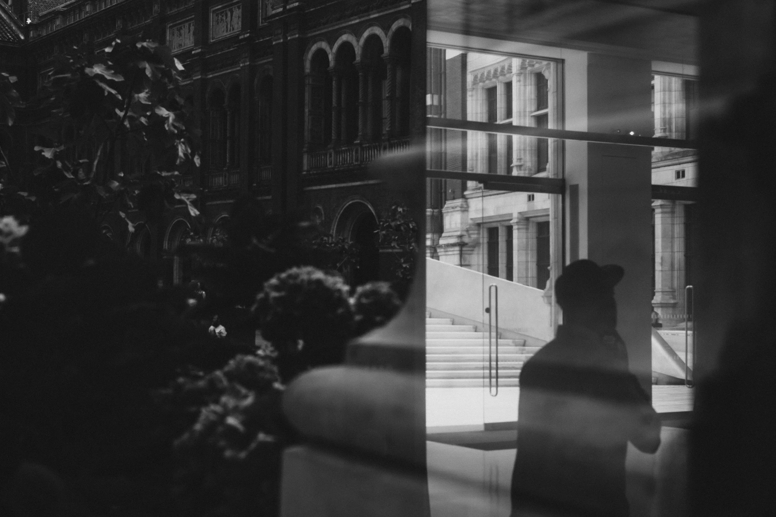 Black and white photo showing a courtyard street view through a glass window, with reflections of a person walking by and buildings on the other side.