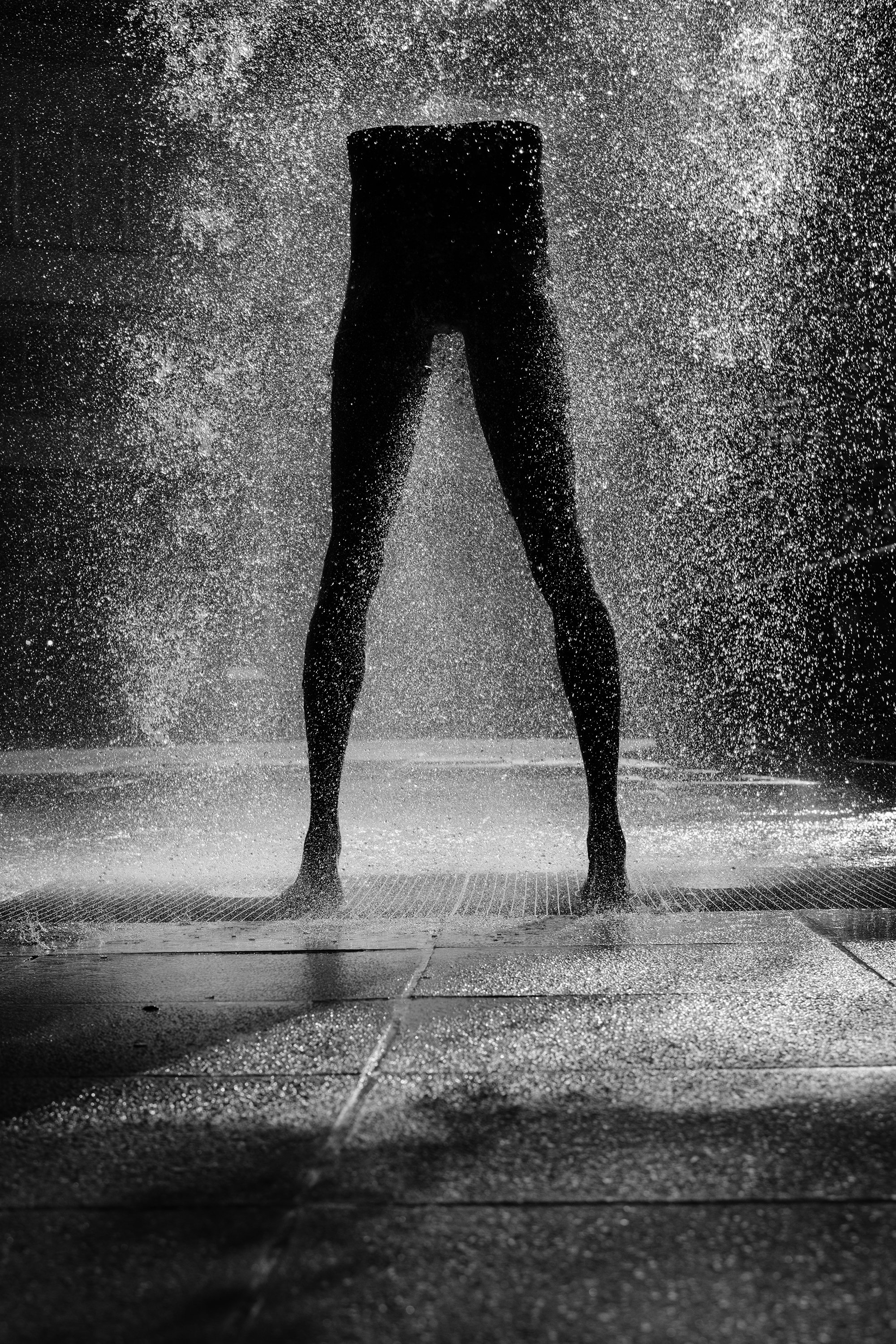 Silhouette of a person standing under a water spray or shower with water droplets illuminated against a dark background.
