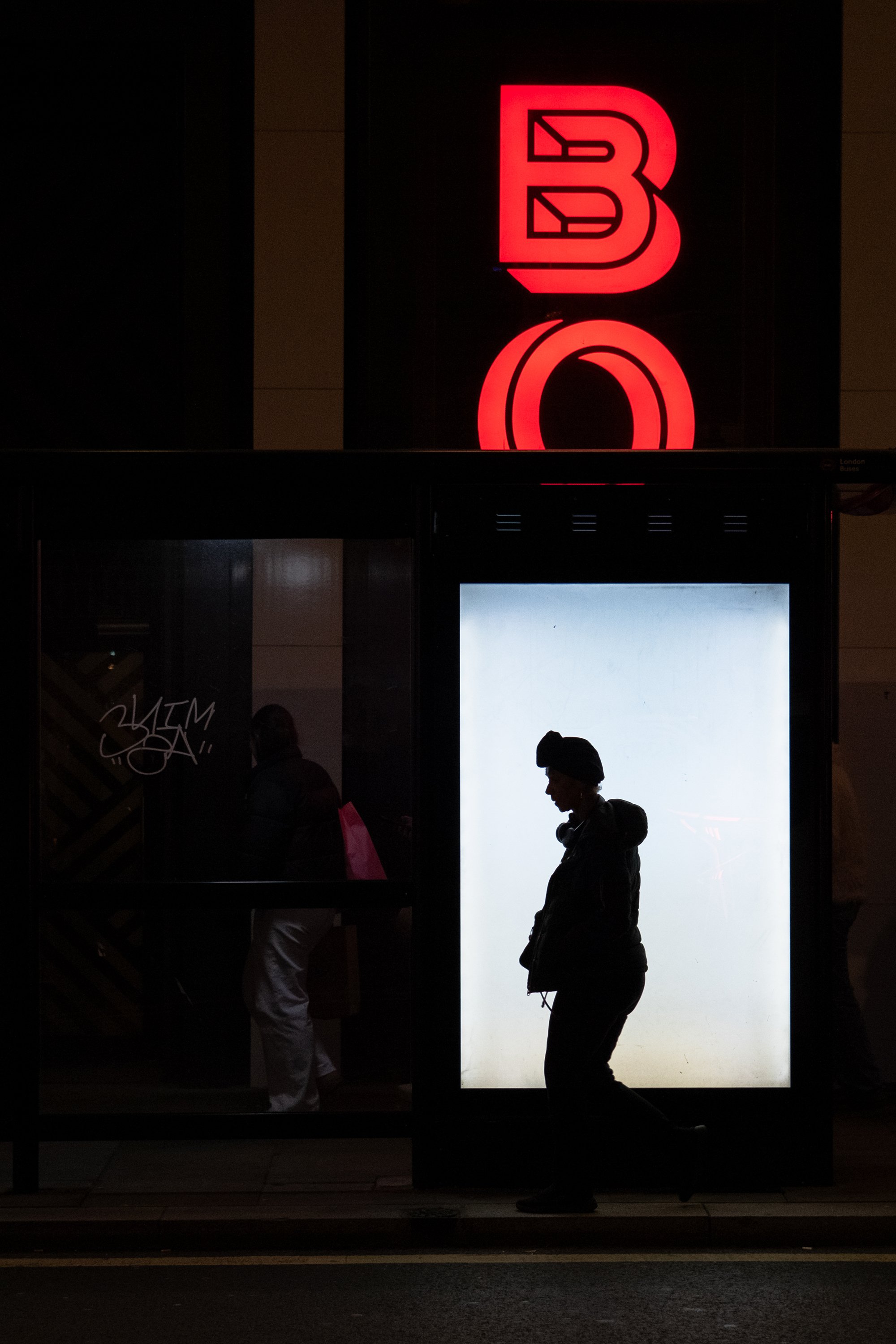 silhouette of a woman walking past a lit bus shelter with a bright white back panel and a red illuminated sign above