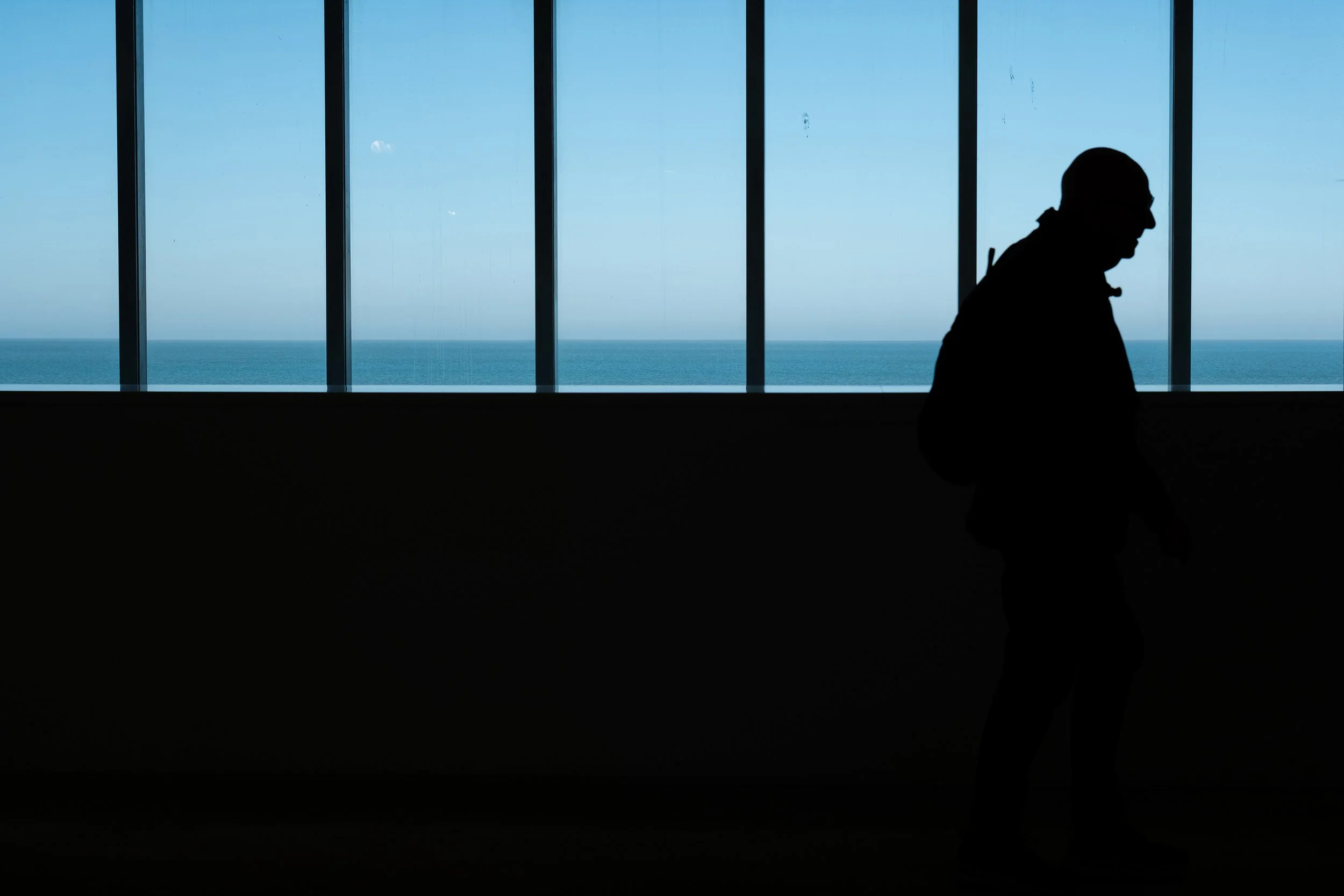 Silhouette of a person walking indoors with a large window showing the ocean and sky outside.