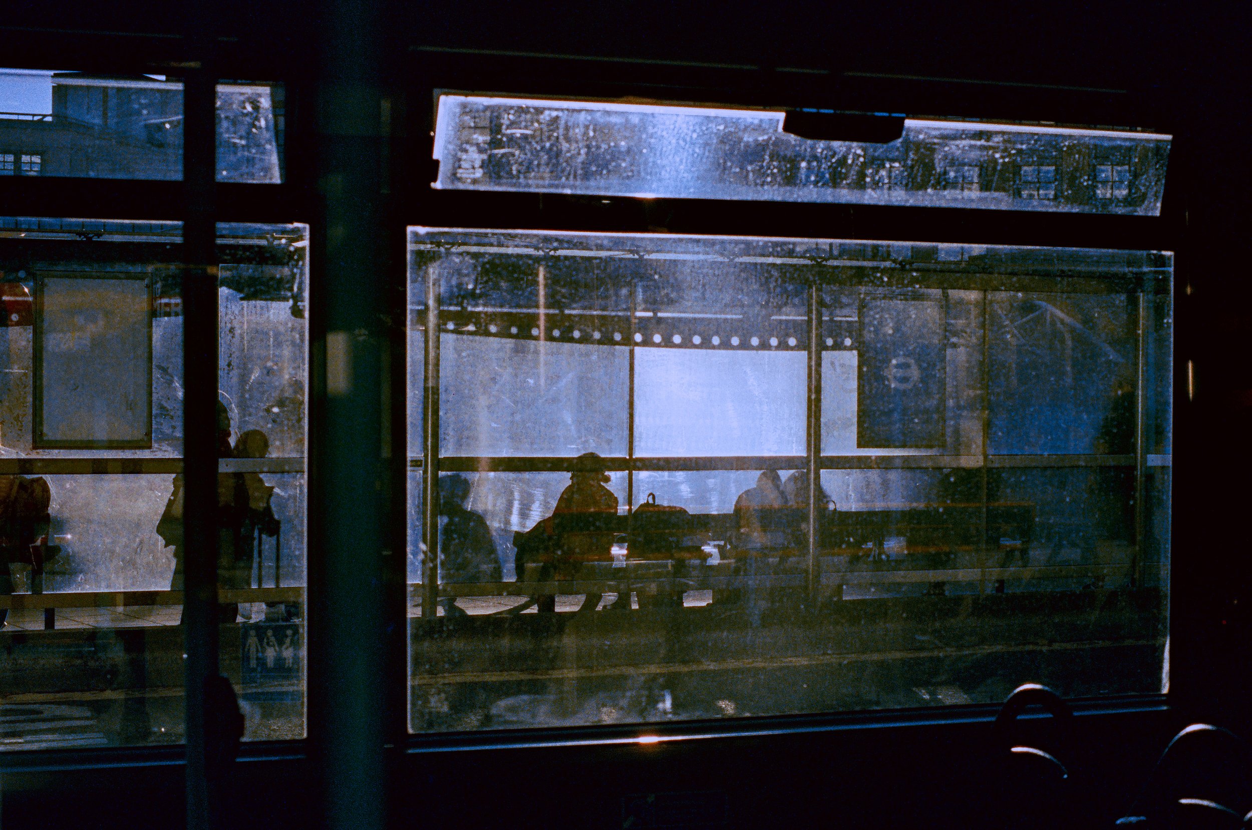 View through a bus window of people waiting at a bus stop. The sunlight creates flares shining through the window. 