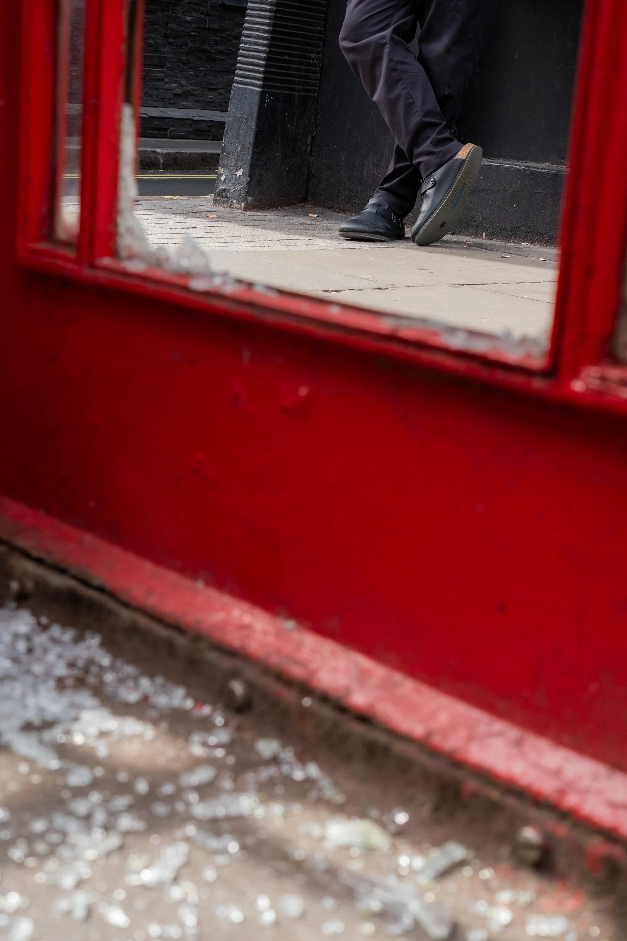 Person wearing dark pants and shoes walking past a red telephone booth on a city sidewalk.