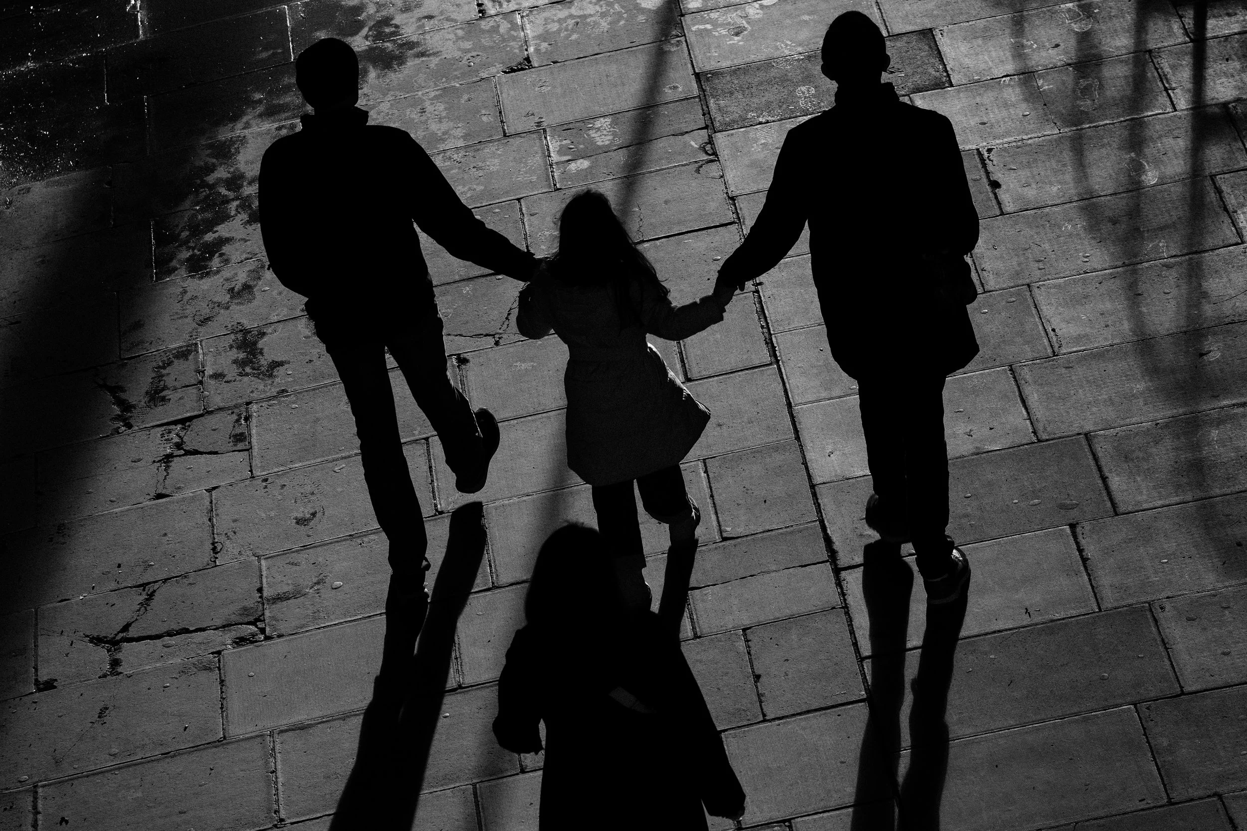 Silhouettes of two adults and a child holding hands and walking on a paved surface. Shadows are cast on the ground in front of them.