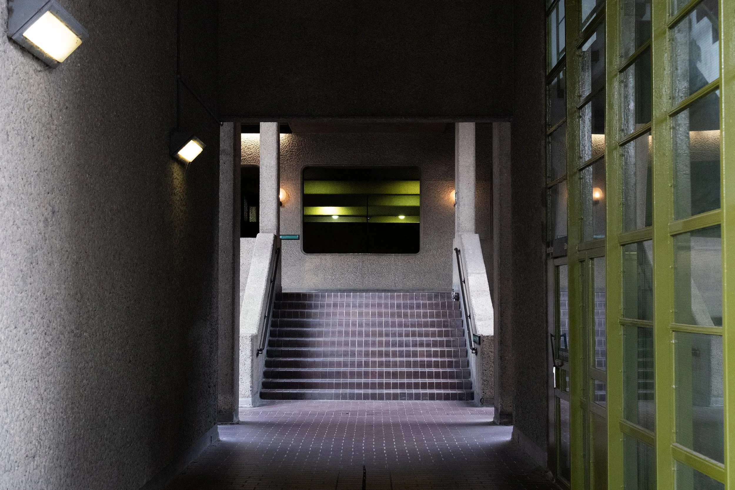 A stairwell with brick steps leading up to a landing, flanked by textured gray walls and illuminated wall-mounted lights, with glass windows and green-colored window frames to the right.