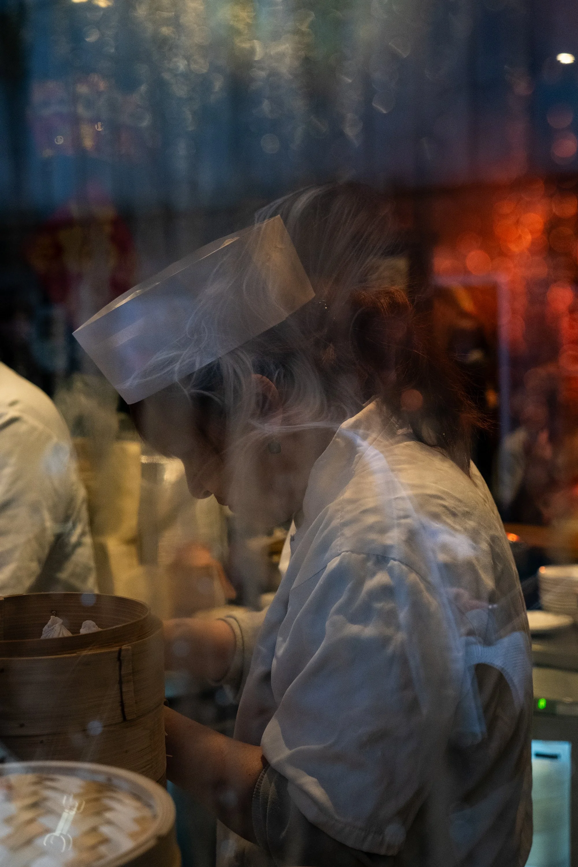 A chef wearing a face shield preparing food in a dimly lit kitchen with steam rising.