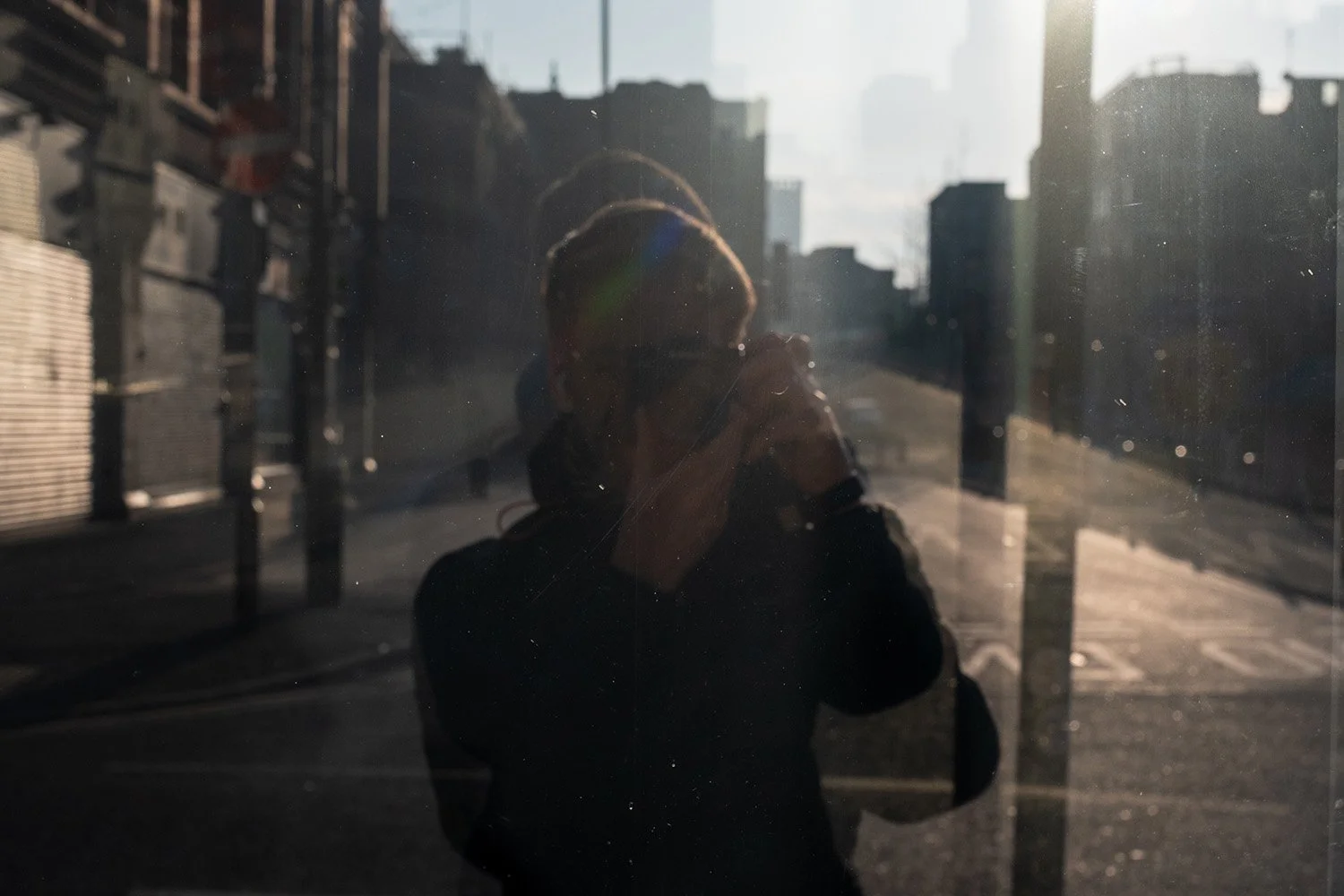 A person taking a selfie through a reflective glass window in an urban area with tall buildings. The person is wearing a dark jacket and sunglasses, with the sun shining brightly behind them.