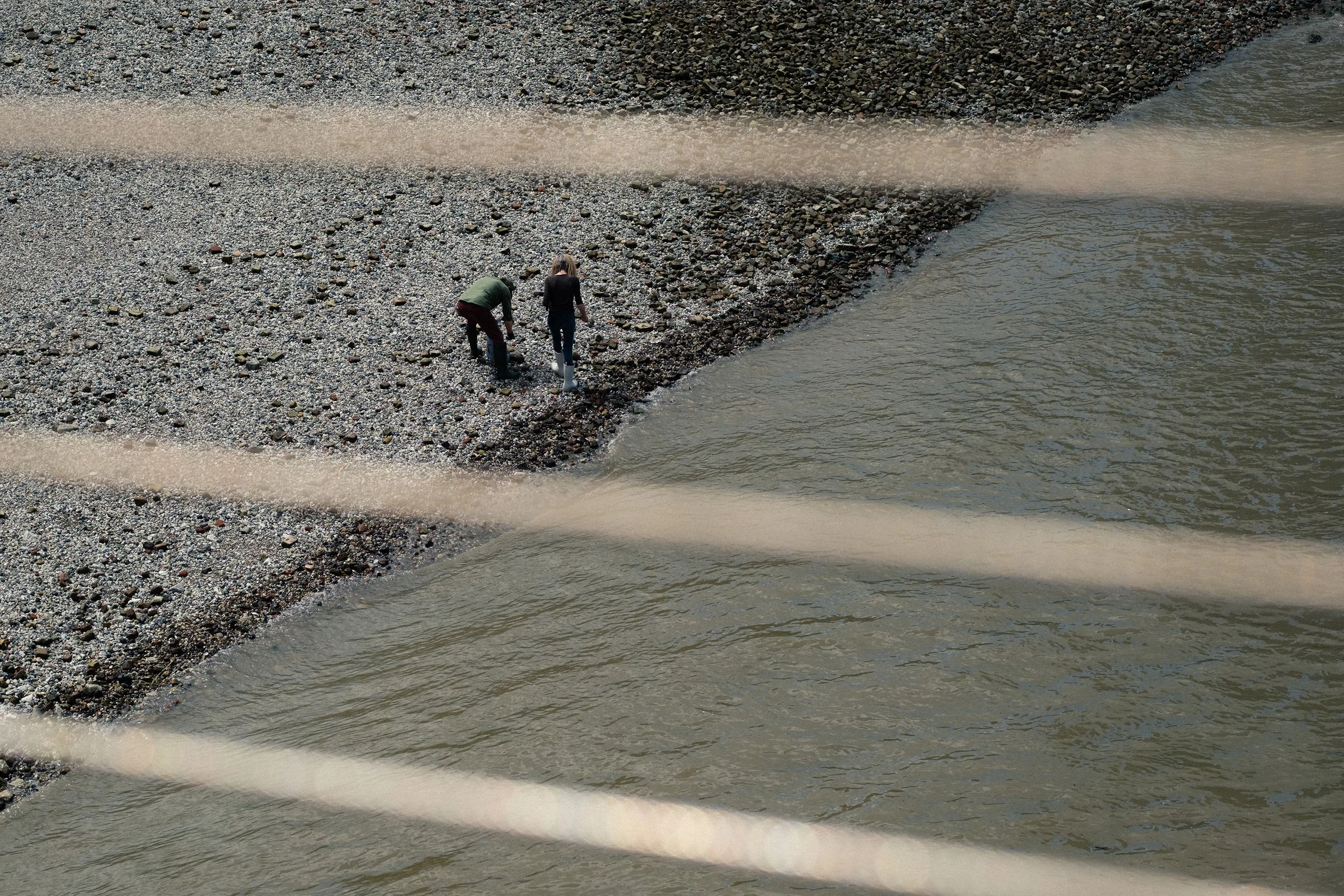 Two people walk along a pebbled shoreline with waves washing up on the shore, seen from above.