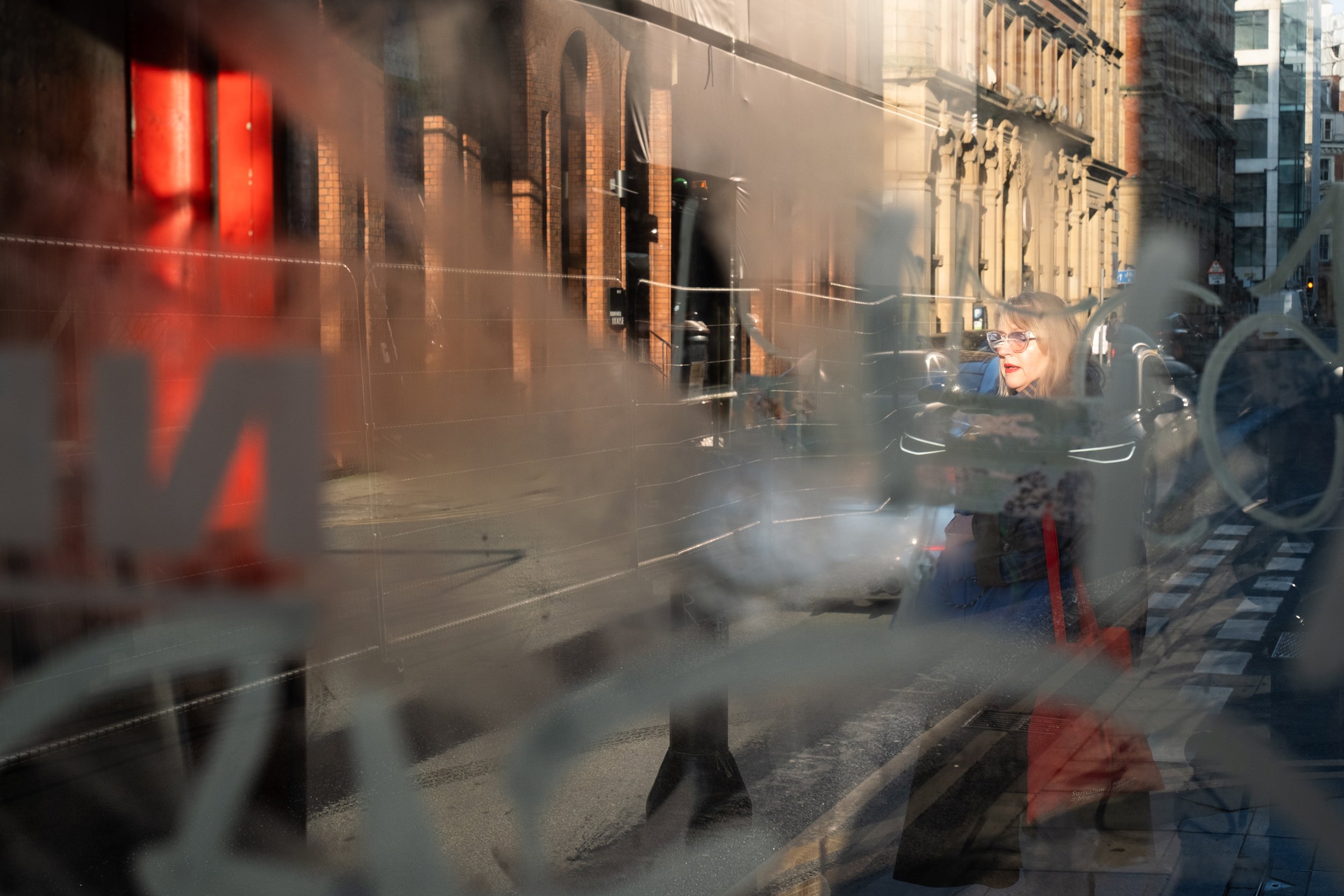 A woman with glasses and blonde hair looking to the right, seen through a foggy or smudged glass window with graffiti, on a city street with brick and stone buildings in the background and steam rising from a street grate.