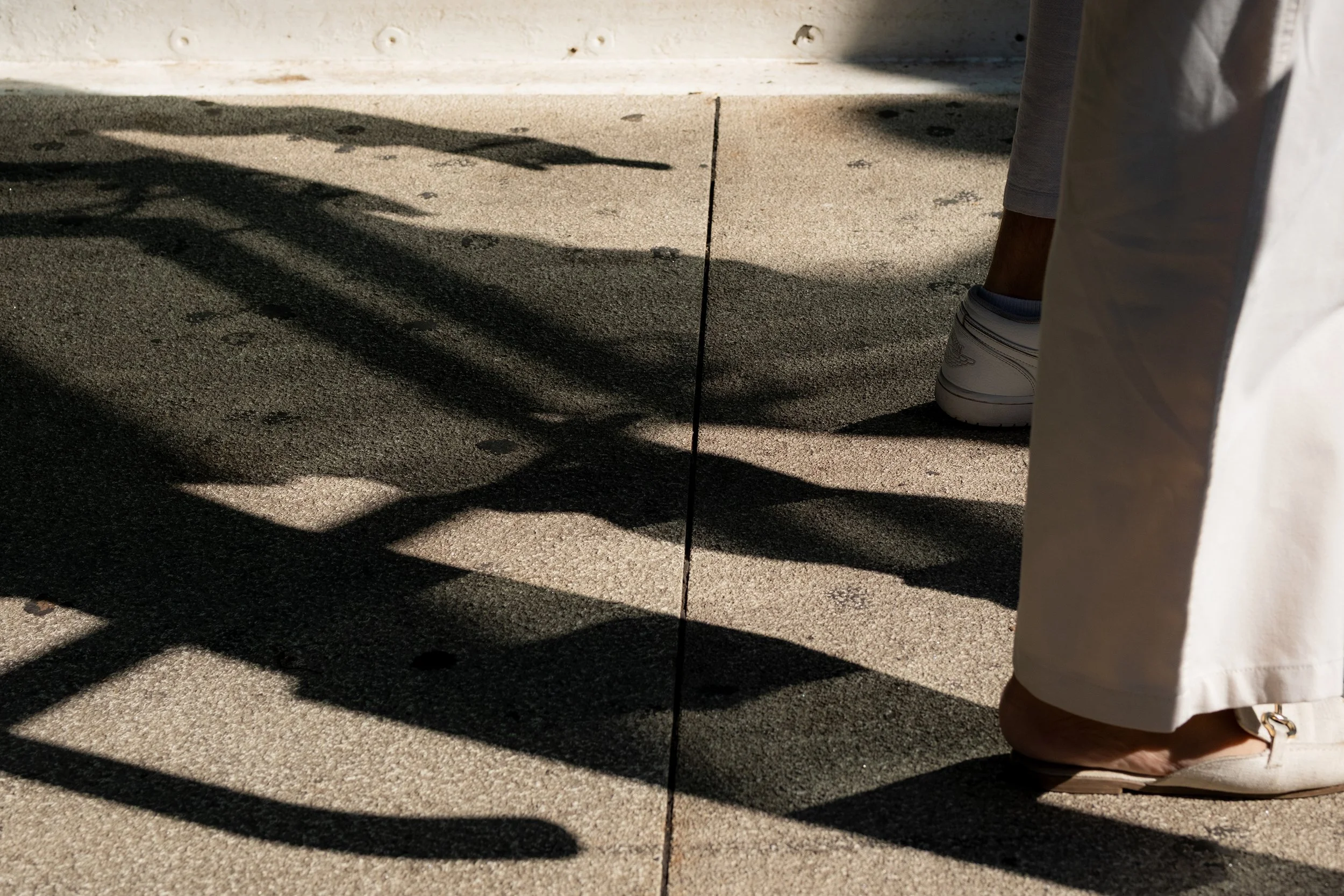A person's legs and feet wearing white shoes and pants, with long white pants covering one foot, standing on a concrete sidewalk. Shadows cast by an object or person are visible on the ground.