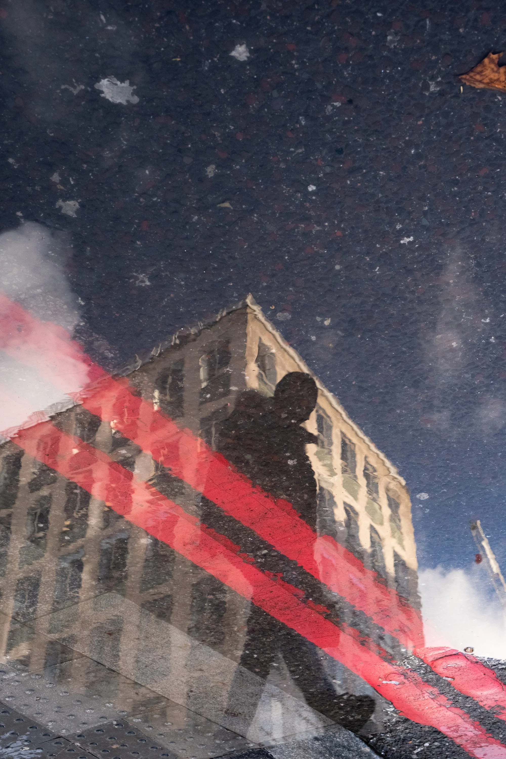 Reflection of a person taking a photo, a building, and a red painted curb on a wet street or sidewalk.