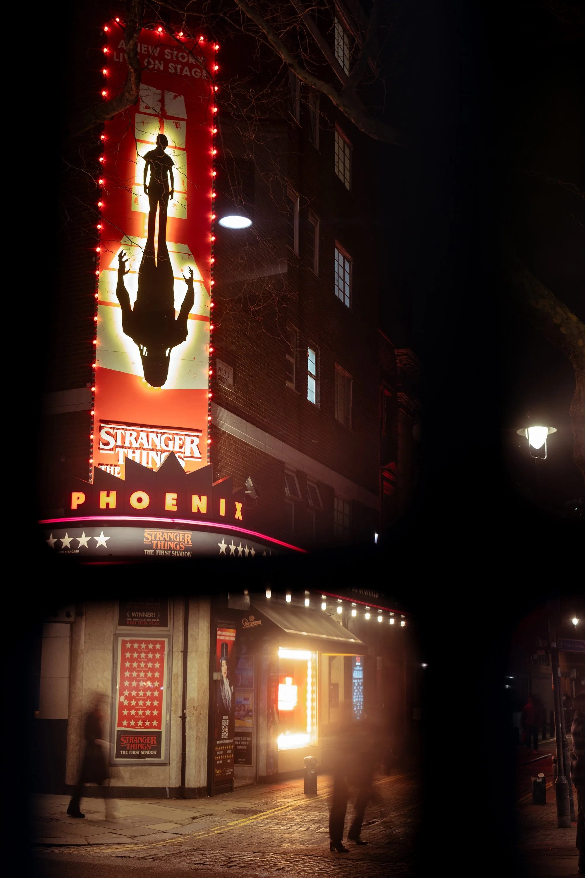 Nighttime street scene featuring a large illuminated vertical poster for the TV series 'Stranger Things,' with the title 'THE FIRST SHADOW' and 'PHOENIX' at the bottom. The poster shows silhouettes of characters and is bordered with red lights. Pedes
