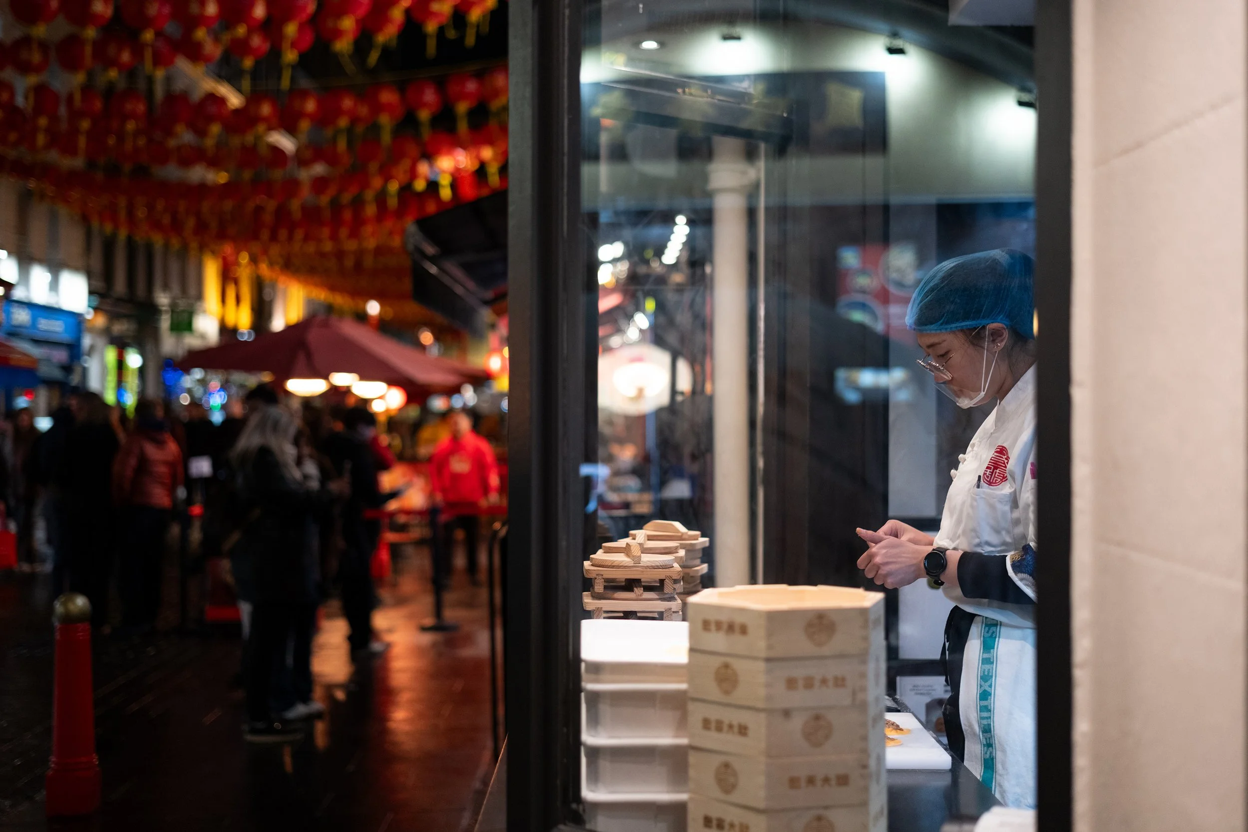 A woman wearing a face mask and hairnet working at a food stand at night in a busy outdoor market. She is preparing or packaging food, with boxes and stacks of food containers in front of her. Blurred people and market lights are visible in the backg