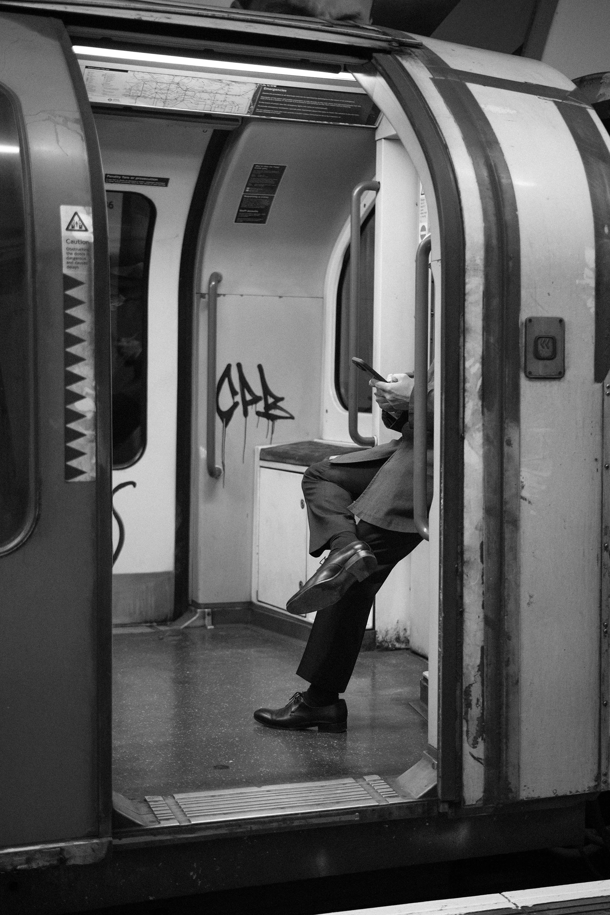 A man in a suit sitting on a tube train, looking at his phone, with graffiti on the wall behind him.