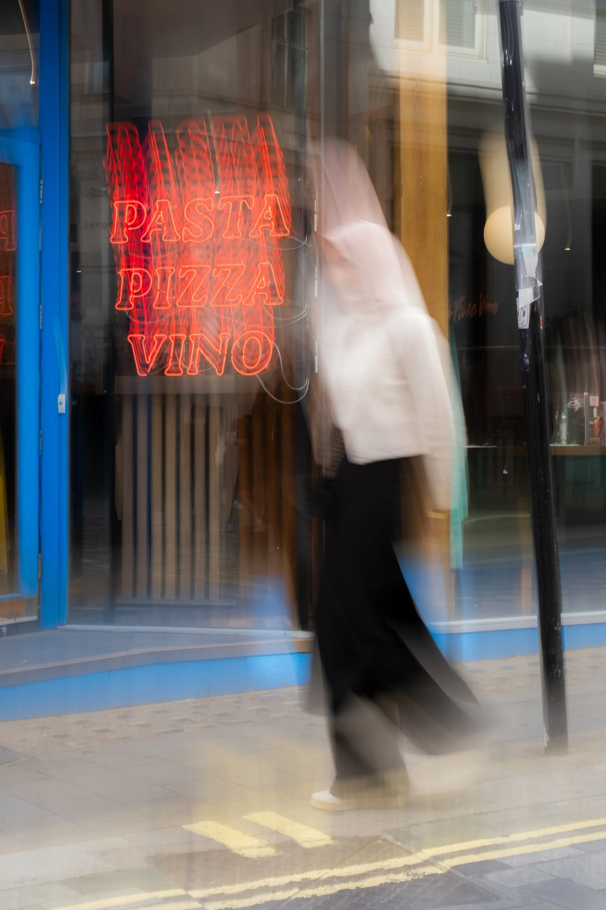 Blurred image of a person walking past a restaurant window with neon signs that read "PASTA," "PIZZA," and "VINO."
