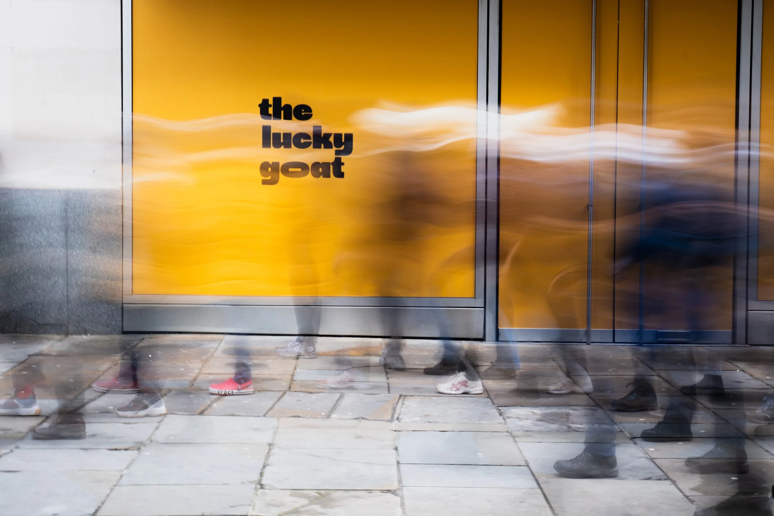 Blurred motion of people walking past a yellow wall with the text 'the lucky goat' written on it, close to an entrance.