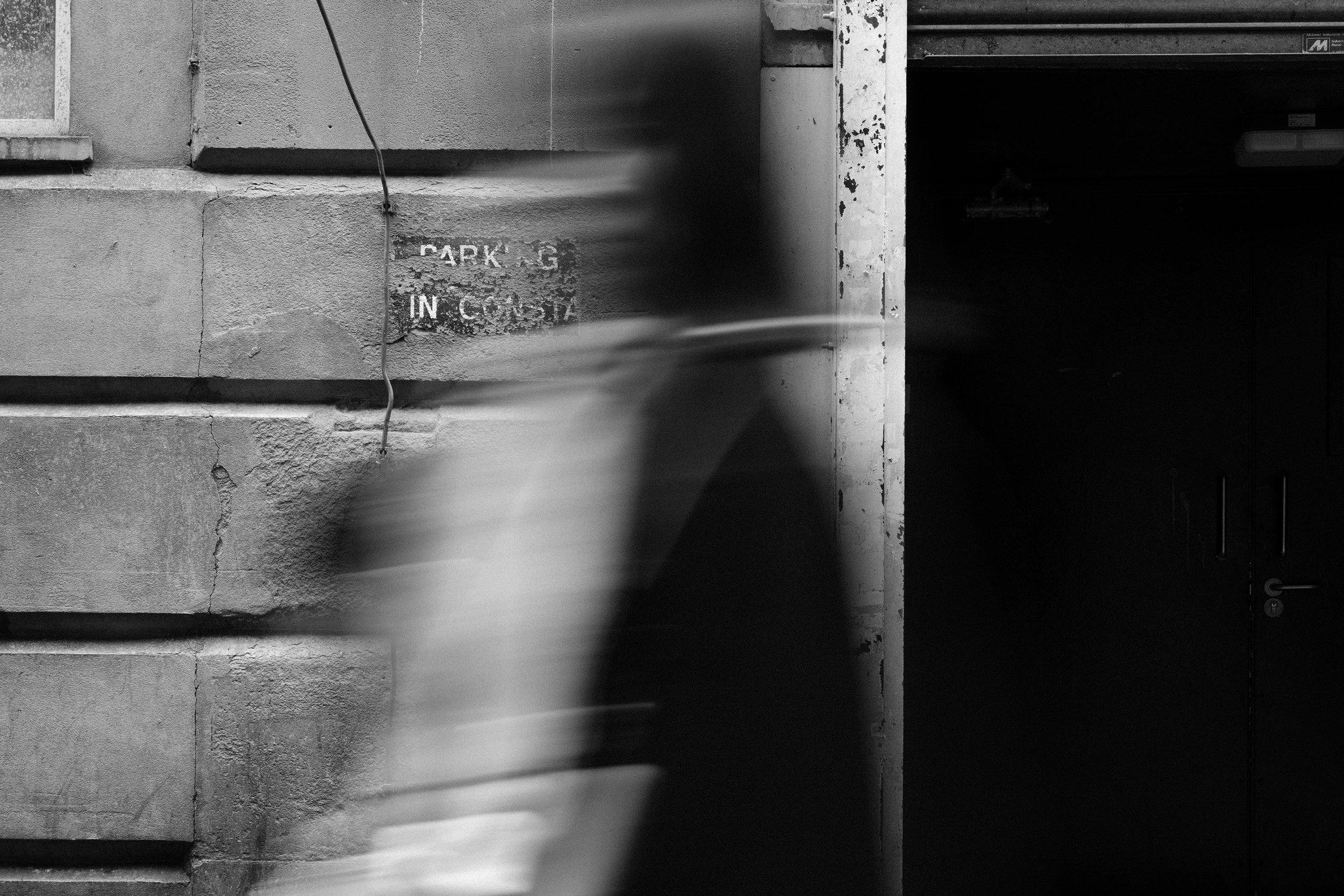 Black and white photograph of an urban alleyway with concrete walls and a dark doorway, featuring a weathered sign that reads 'PARKING IN C' and a vertical pipe on the left side.