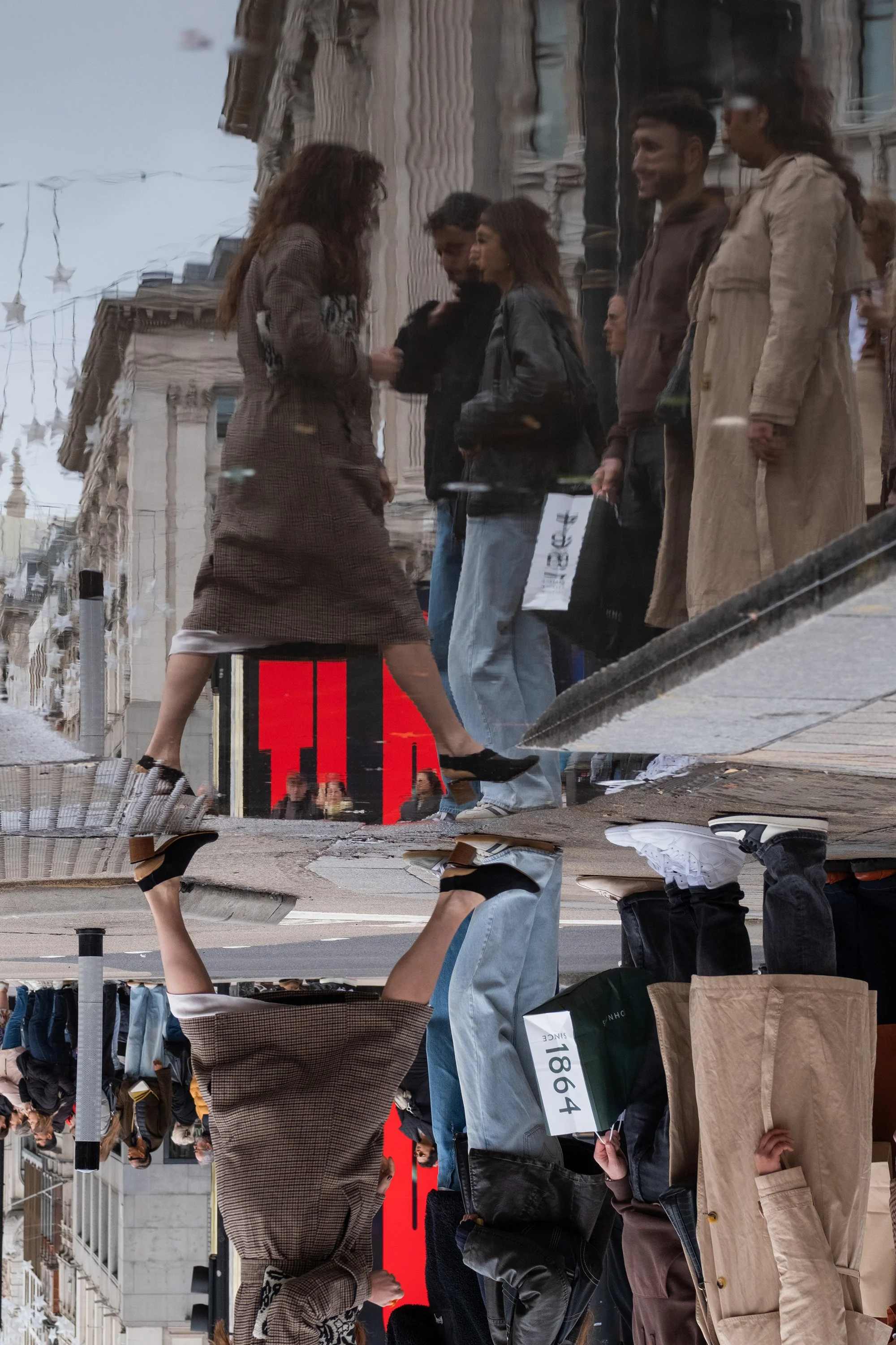 Reflection of people walking and talking on a city street with buildings and a large red sign in the background, captured in a puddle or shiny surface.