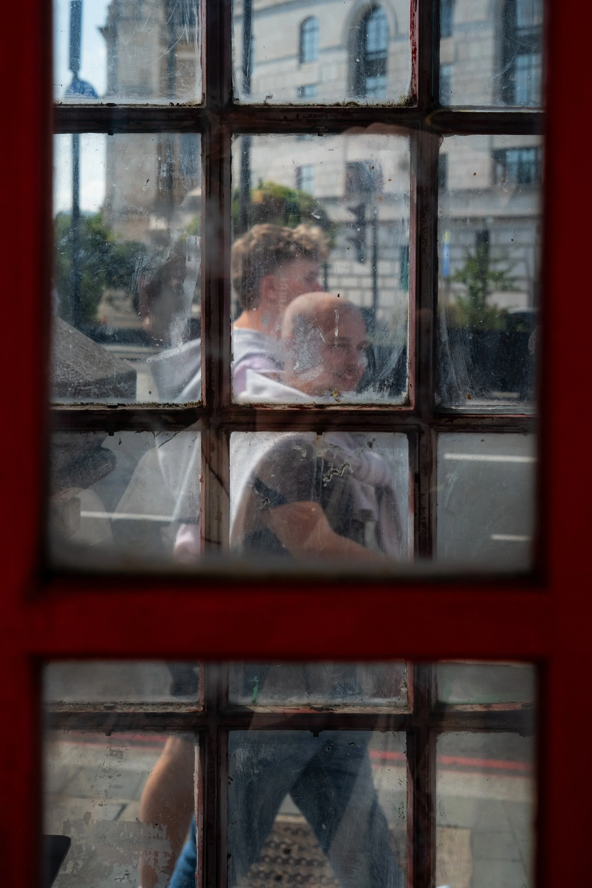 Two people walking outside through the glass of a red phone booth. The window appears old and has dirt and grime on it. In the background, buildings and street are visible.