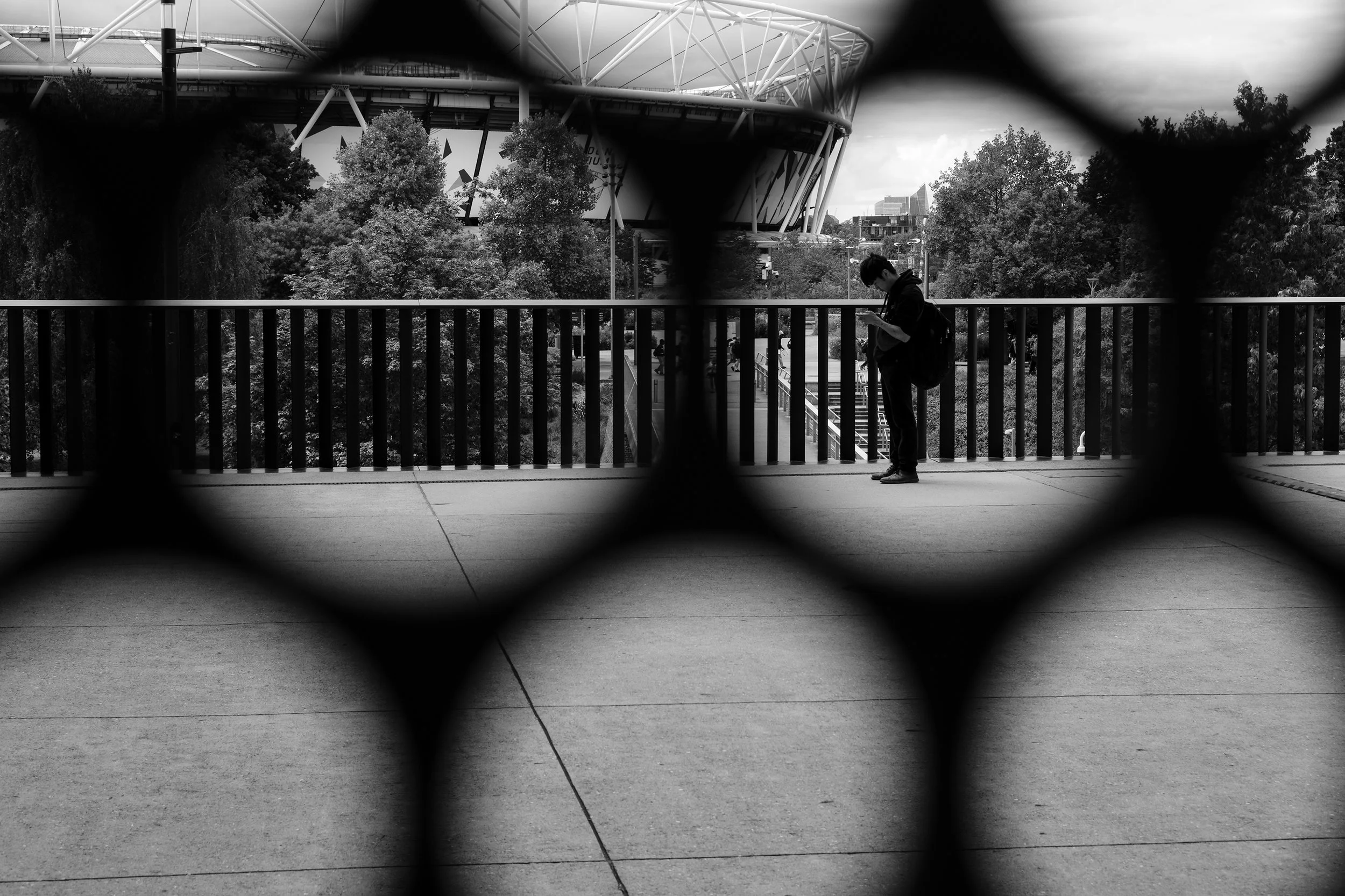 A person looking at their phone, seen through a metal railing with a circular pattern, with trees and a modern stadium in the background.