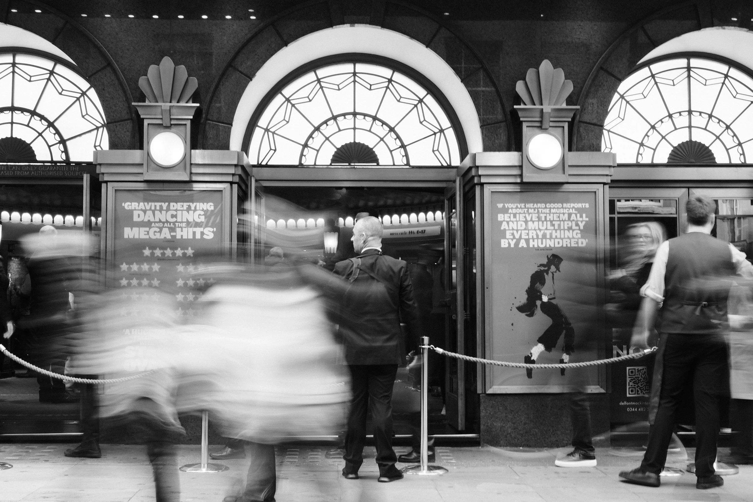People entering a theater entrance with posters advertising a musical, with blurred motion of pedestrians outside and a man in a suit standing near the entrance.