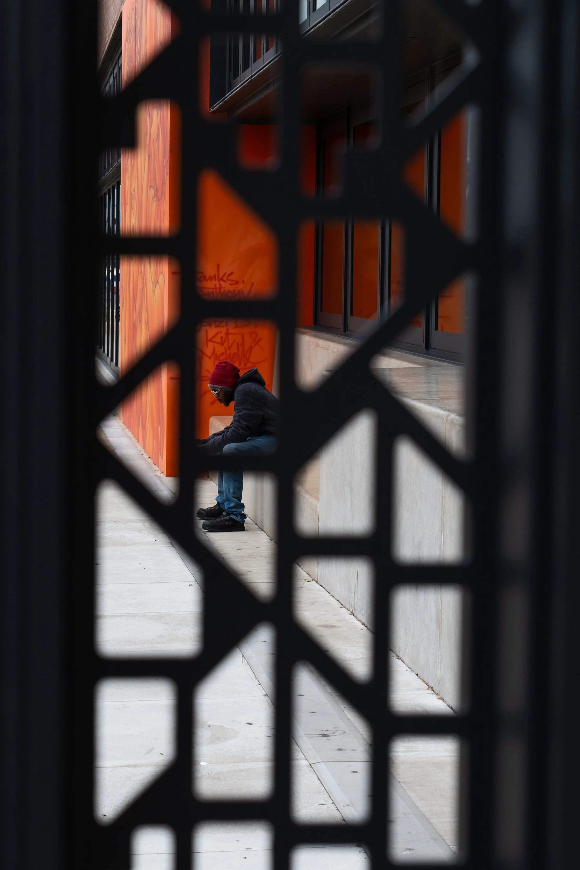 A person in a red hat and sunglasses and glasses sitting on a bench, seen through a black decorative metal fence, with a red wall in the background.