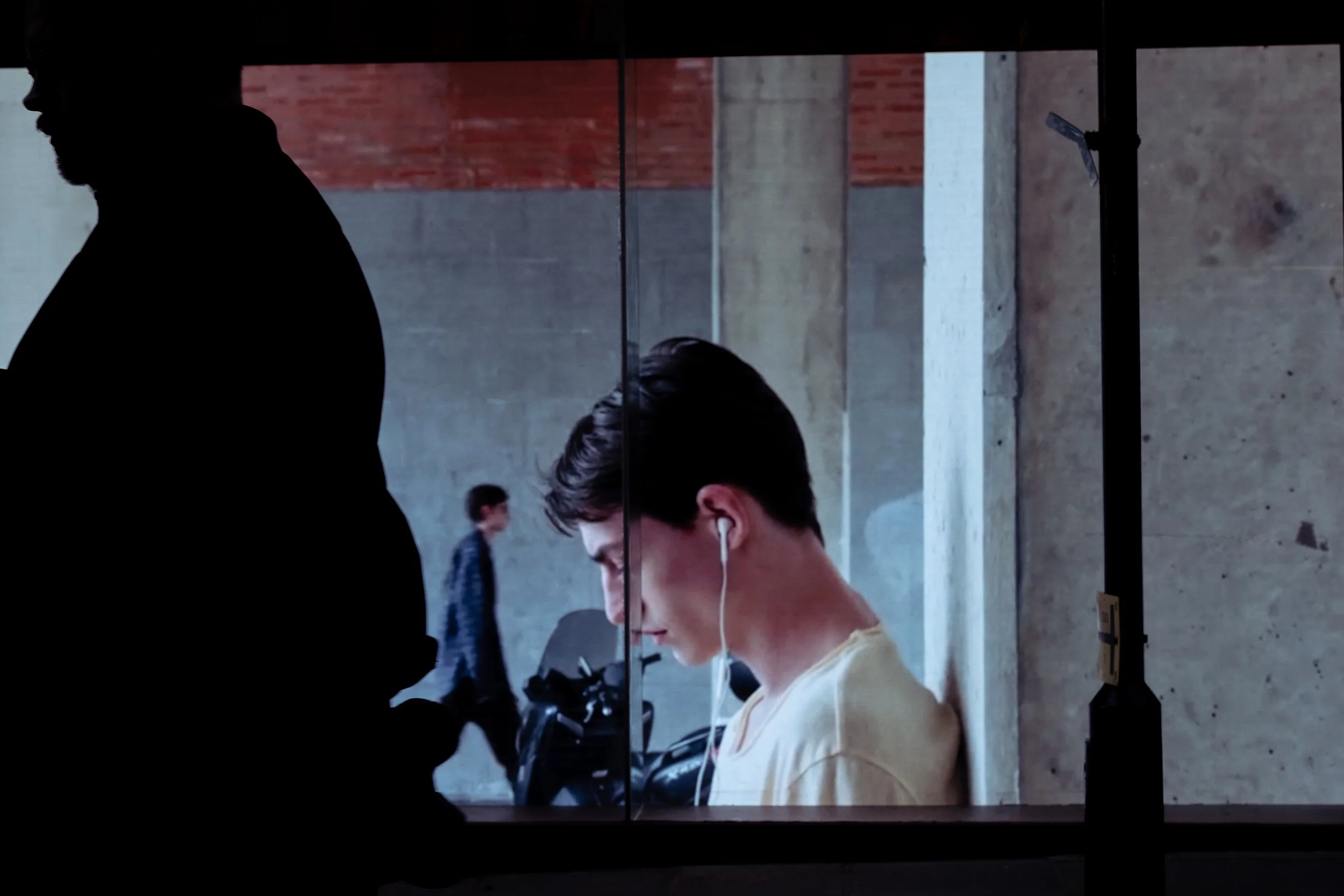 Silhouetted man walking past a large LED screen showing a young man wearing earphones, focused on his device, in an urban environment with concrete and brick walls.